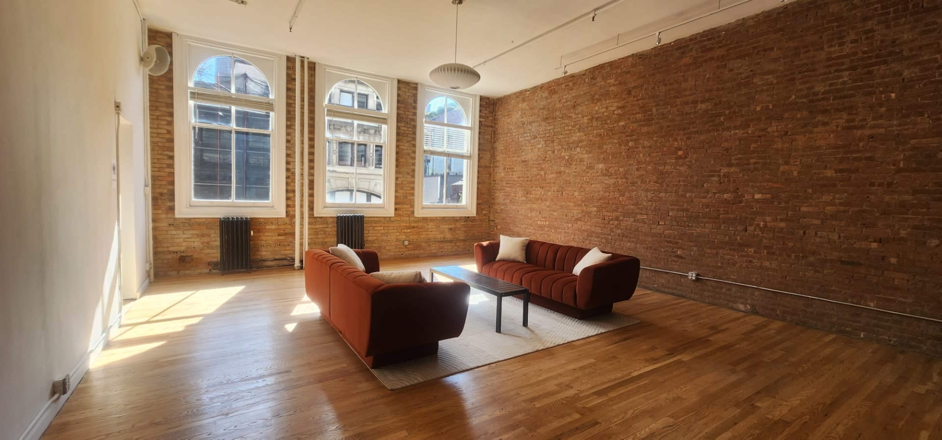The image shows a spacious room with exposed brick walls, featuring two orange couches and a coffee table on a light-colored rug, illuminated by natural light from large windows.