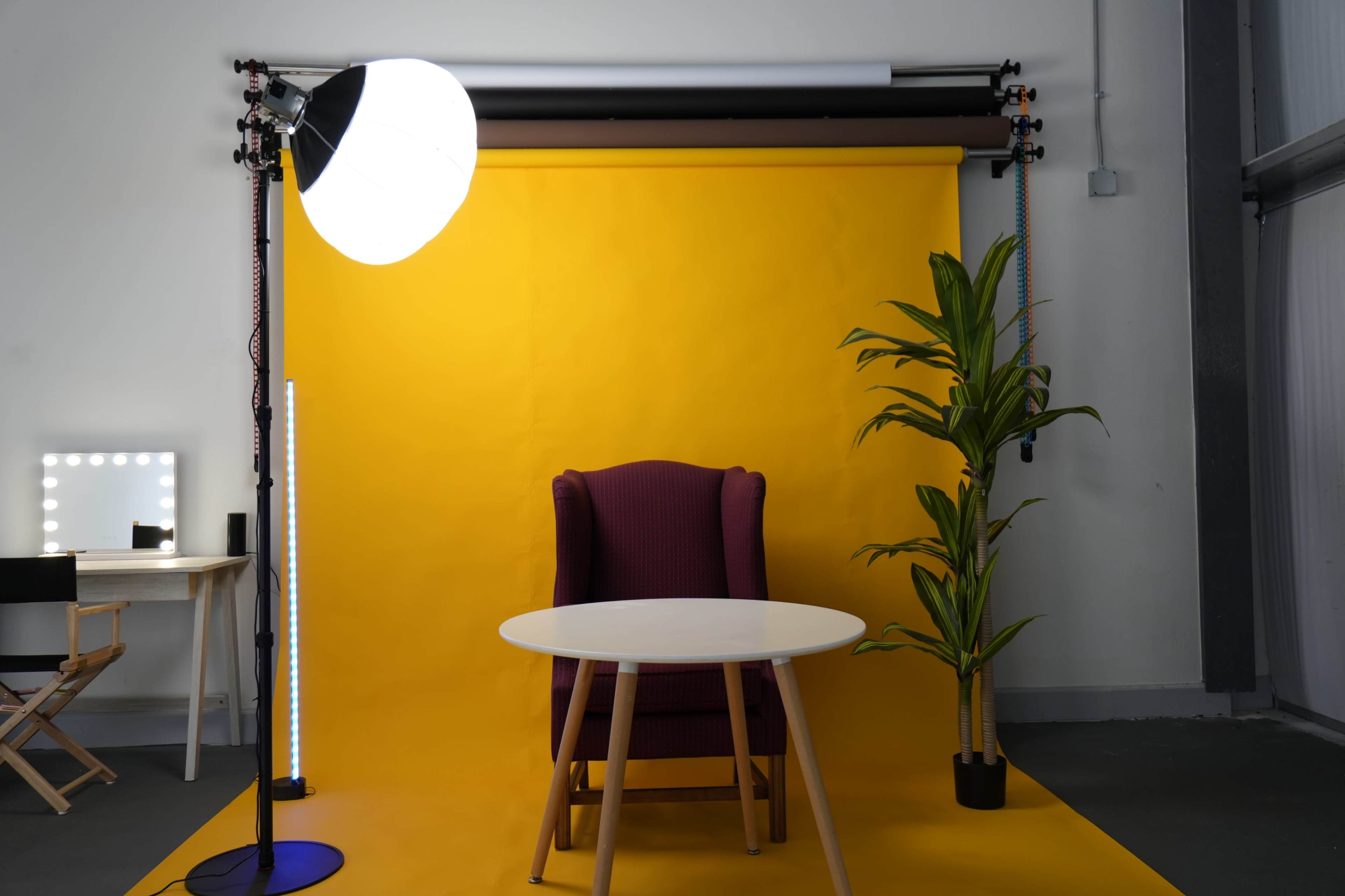 The image features a brightly lit studio setup with a yellow backdrop, a purple upholstered chair, a round white table, and a potted plant.