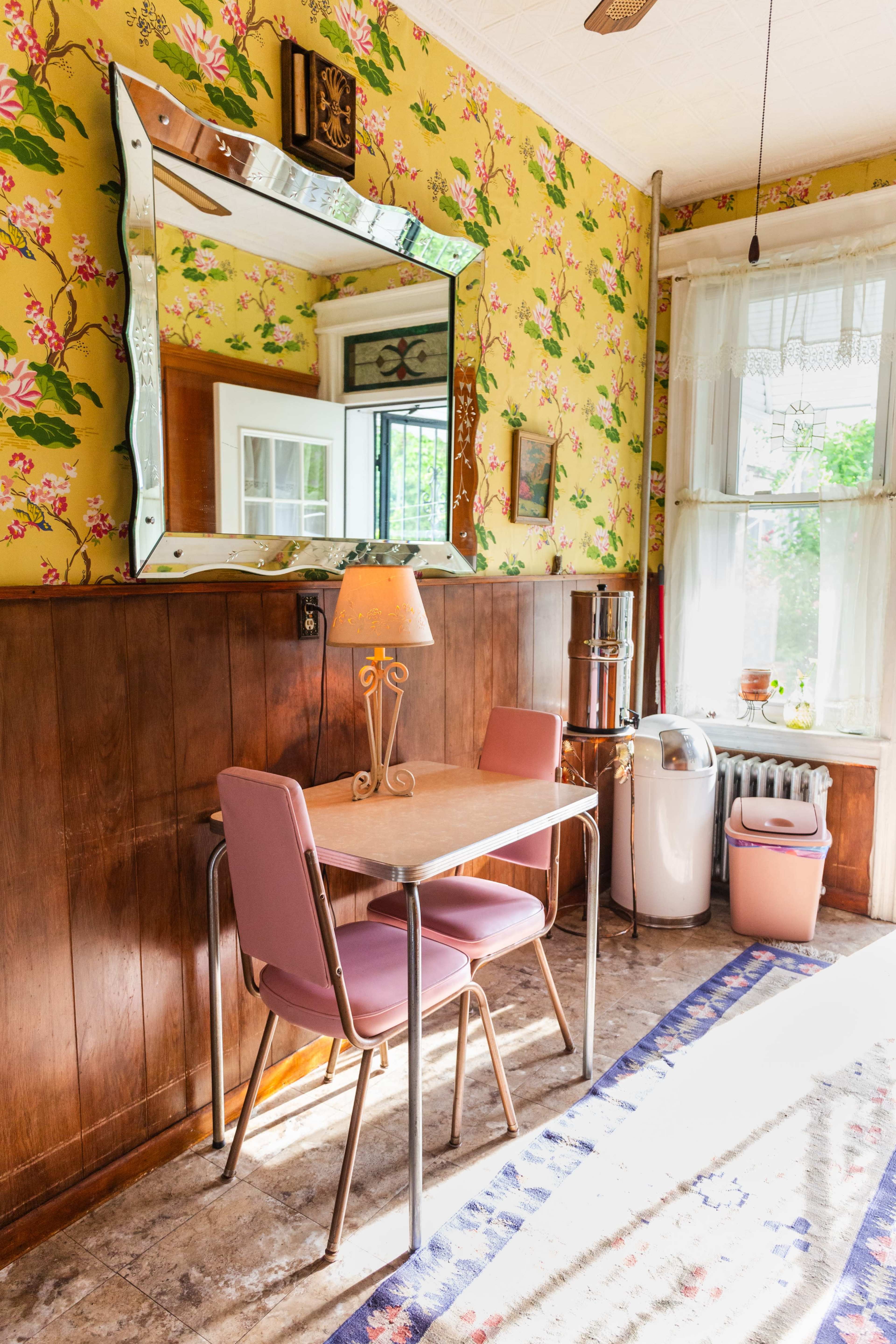 The image shows a bright room with floral wallpaper, a wooden feature wall, a small table with two pink chairs, and various furnishings including a lamp and a radiator.