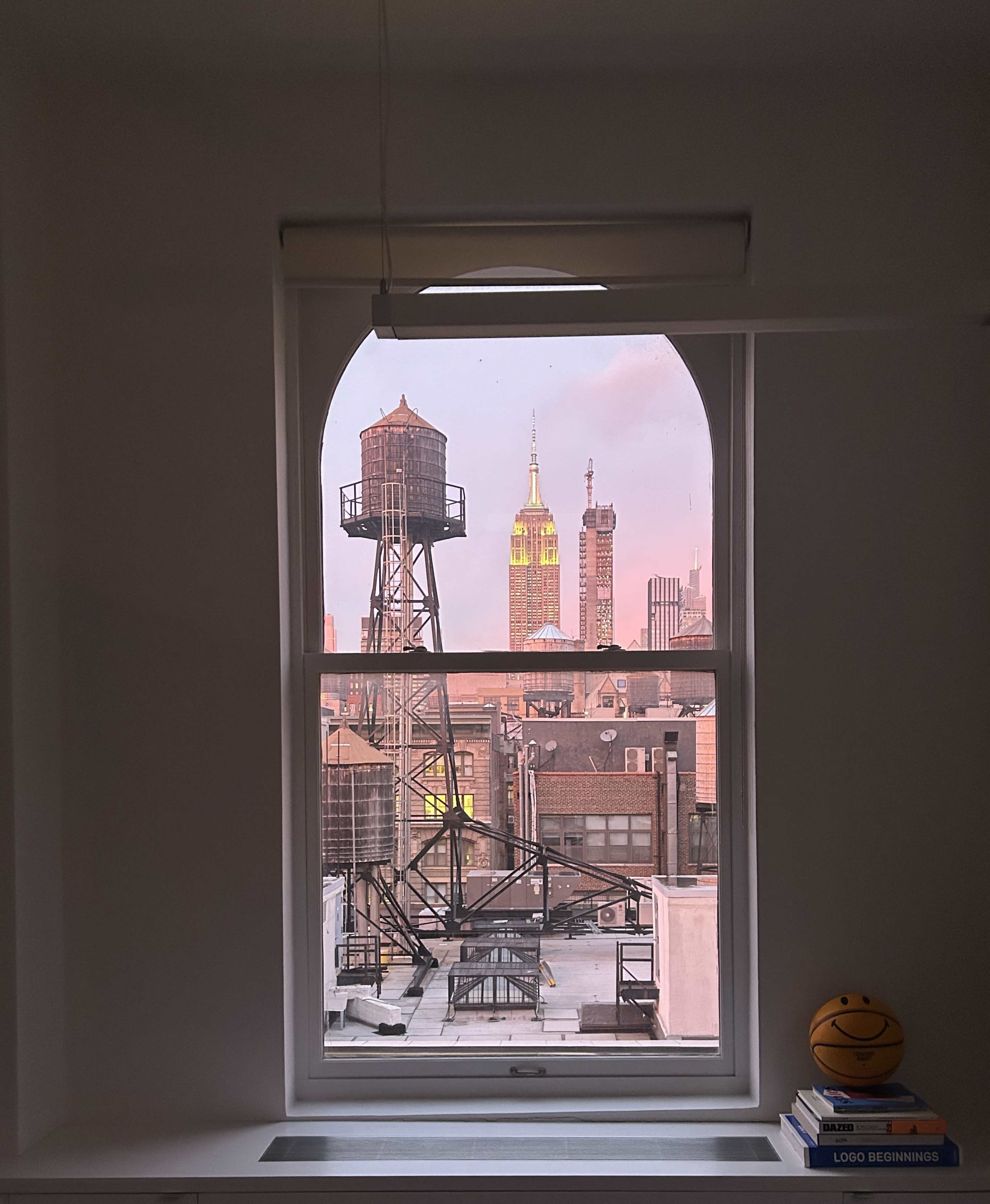 The view from the window shows the Empire State Building alongside a water tower and various rooftops during sunset.
