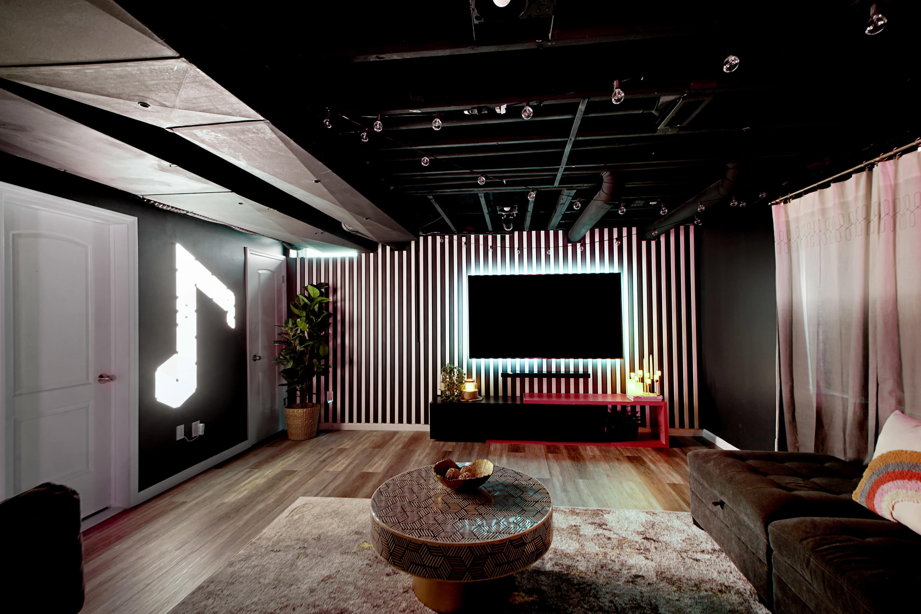 The interior of a modern living room features a large wall-mounted TV, decorative lighting, and a plush couch with a round coffee table in the center.