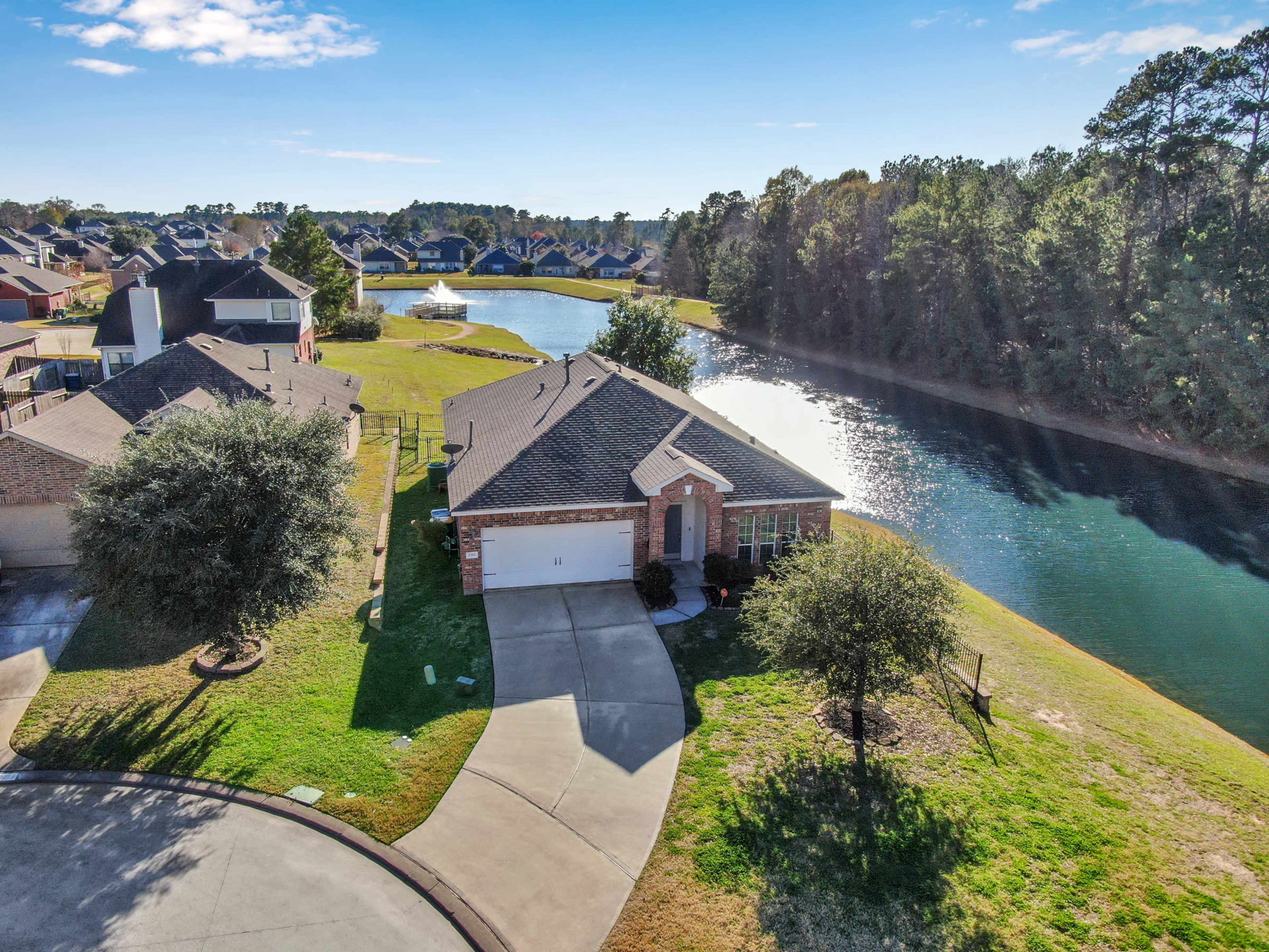 The image shows a single-story brick house situated near a winding waterway, surrounded by lush greenery and residential homes.