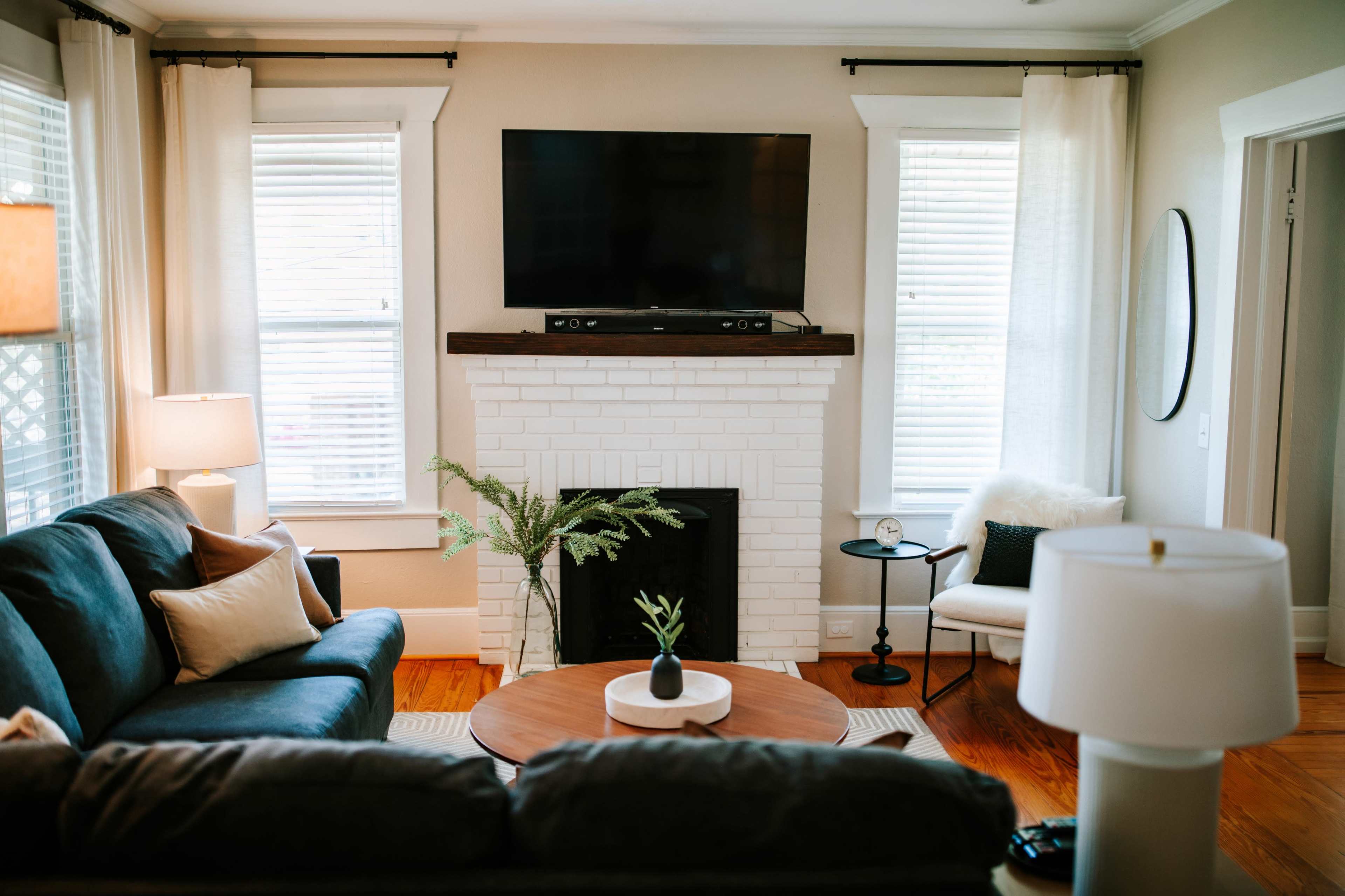 The image shows a cozy living room featuring a dark blue sofa, a round coffee table, and a white brick fireplace with a television mounted above it.