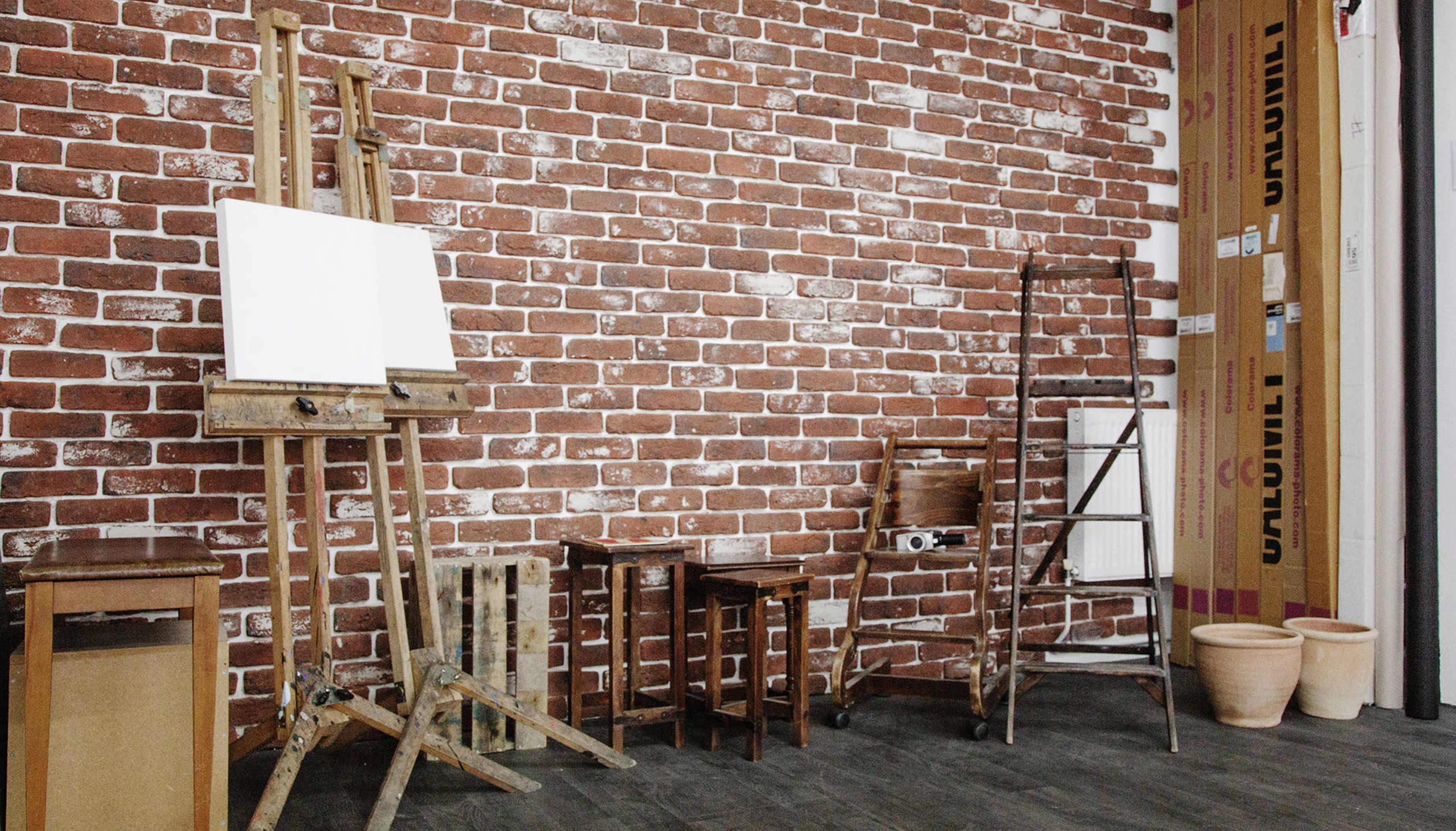 An interior space with a brick wall, an easel and two wooden tables, along with a ladder and two empty flower pots.