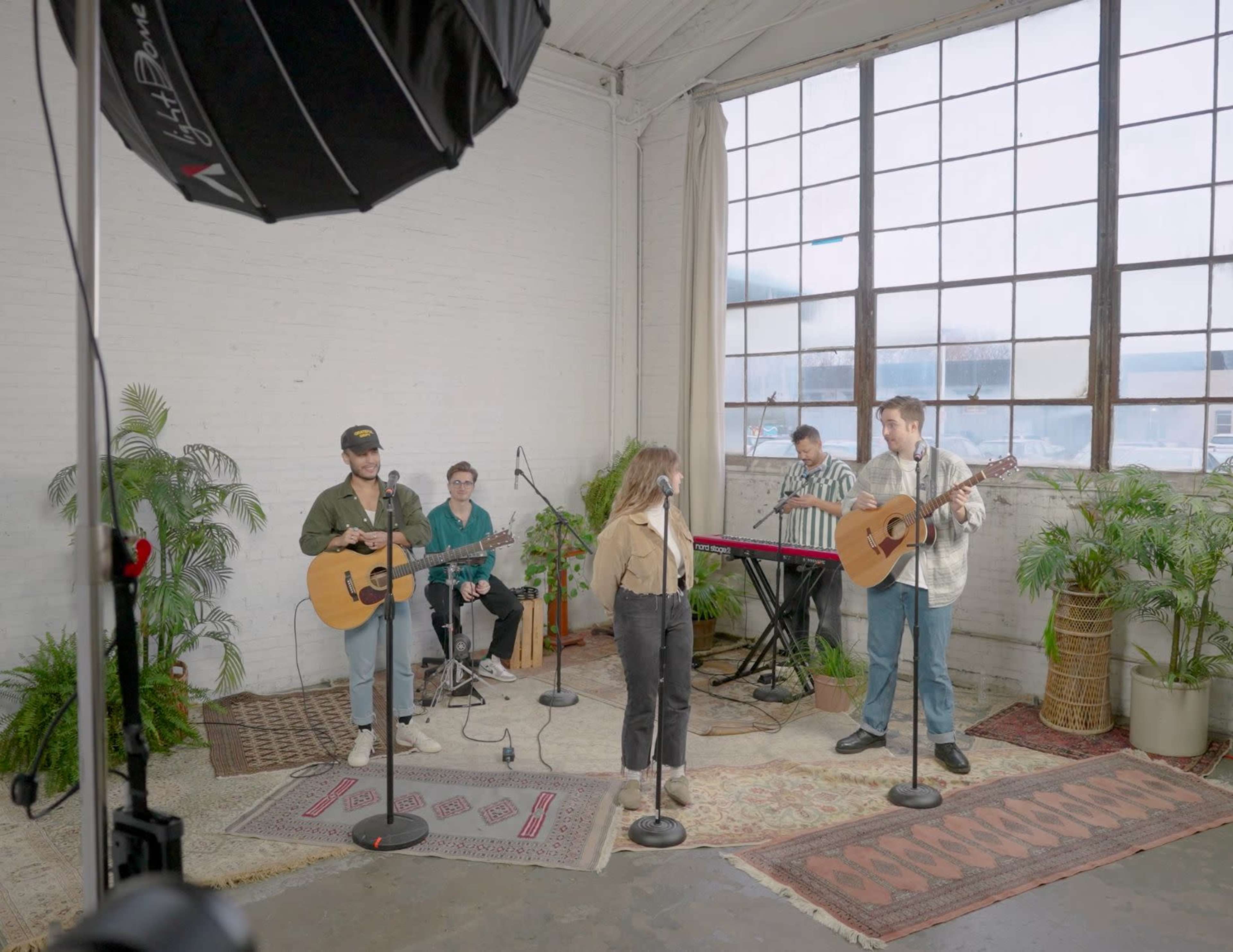 A group of musicians is performing with guitars and keyboards in a studio space featuring large windows and potted plants.