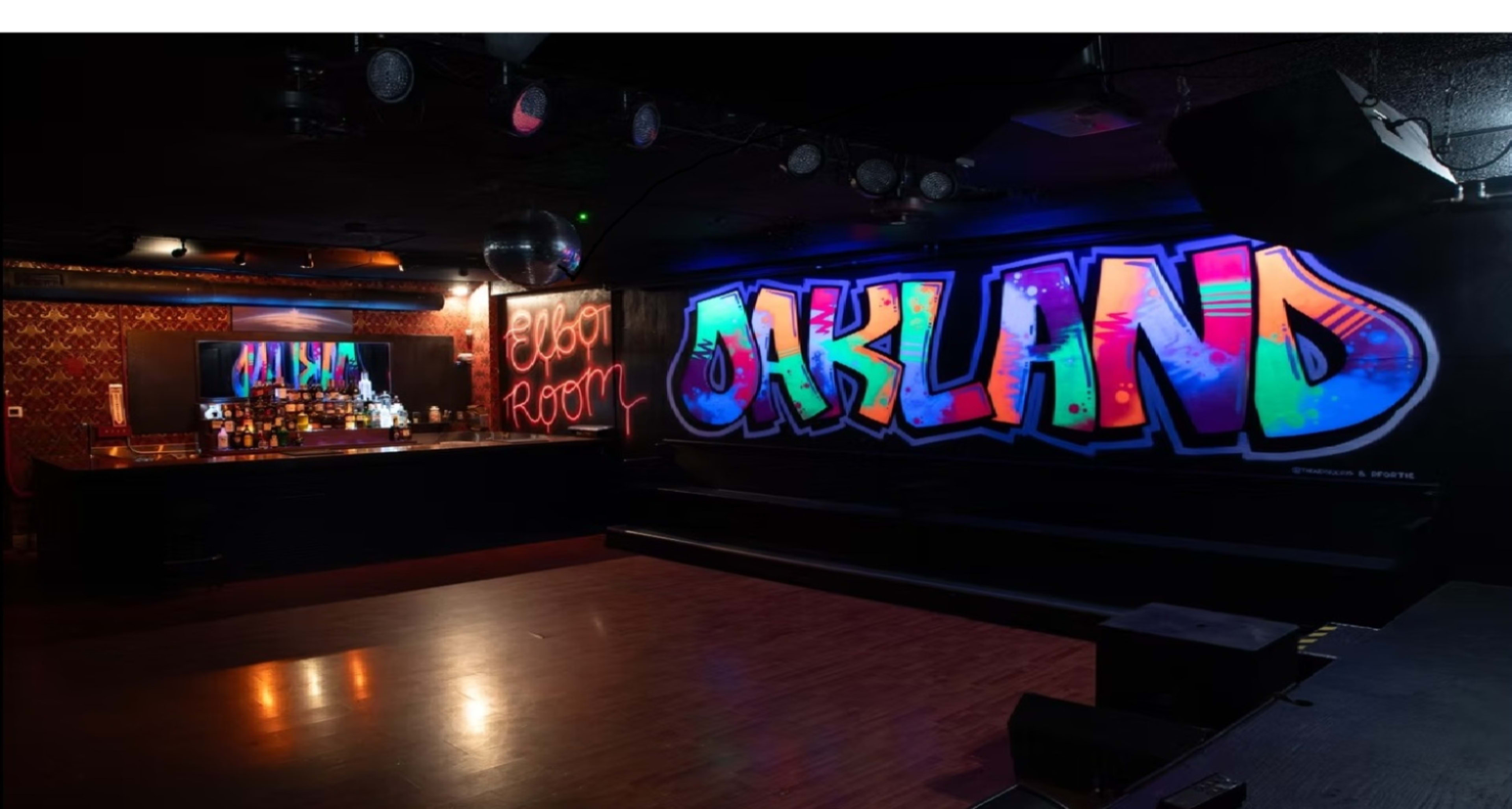 The image shows a dimly lit bar area featuring a colorful graffiti mural of the word "OAKLAND" on a wall, with a bar counter and mirrors visible in the background.