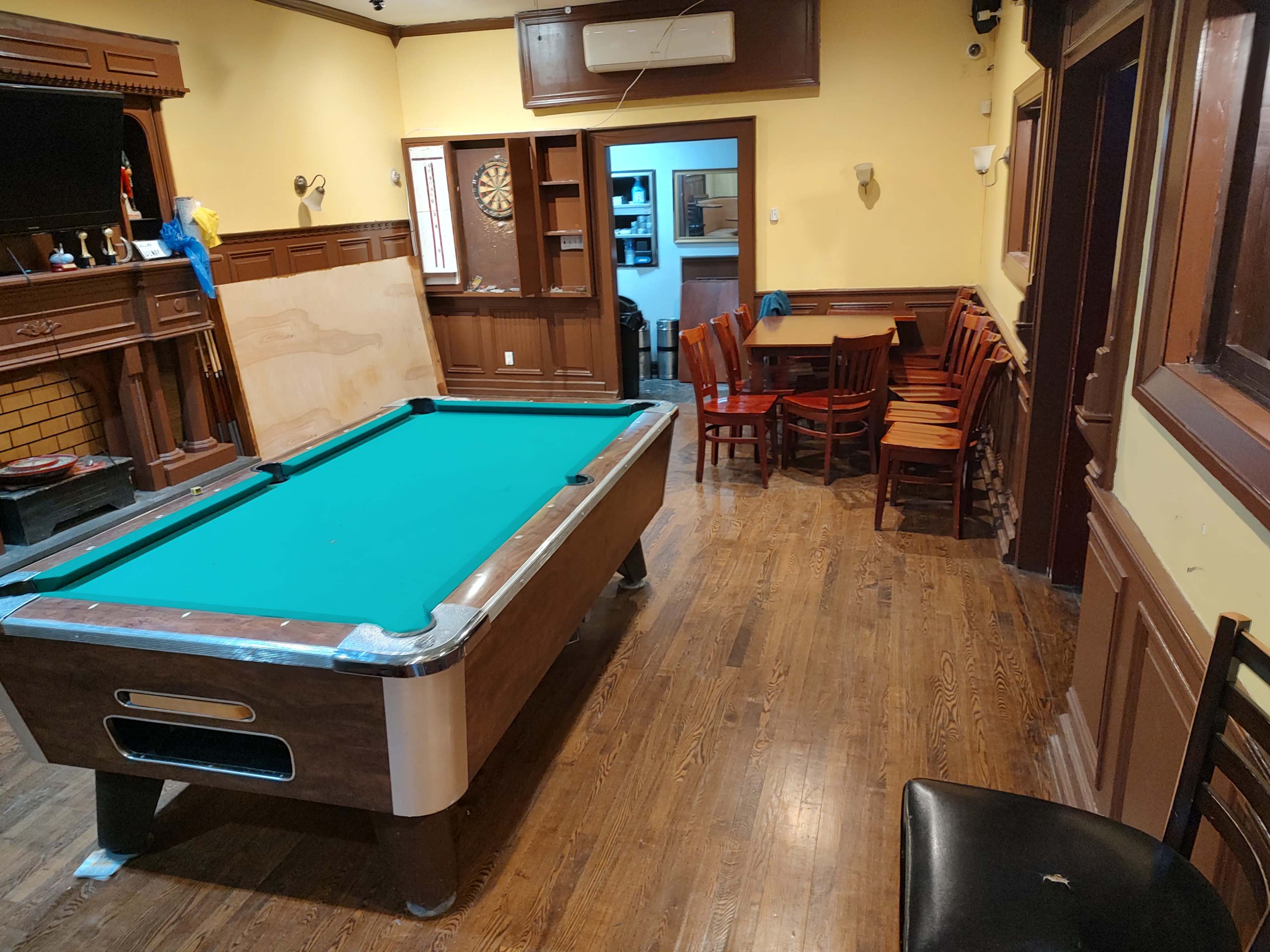 The image shows a room with a green-felt pool table on a wooden floor, surrounded by wooden paneling, and featuring a seating area with several chairs and a table in the background.