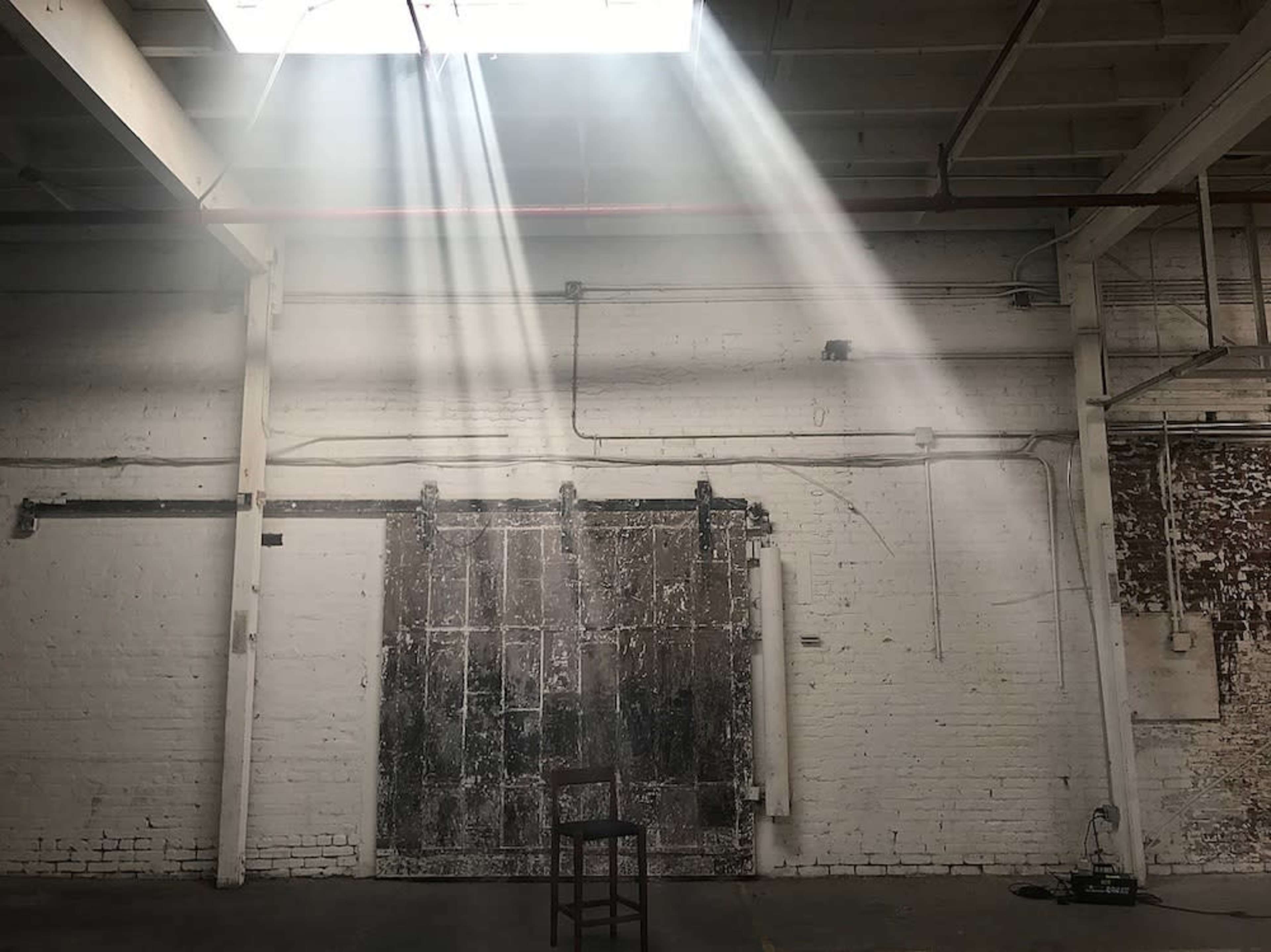 A single wooden chair is positioned in an empty industrial space illuminated by beams of light streaming through a skylight.