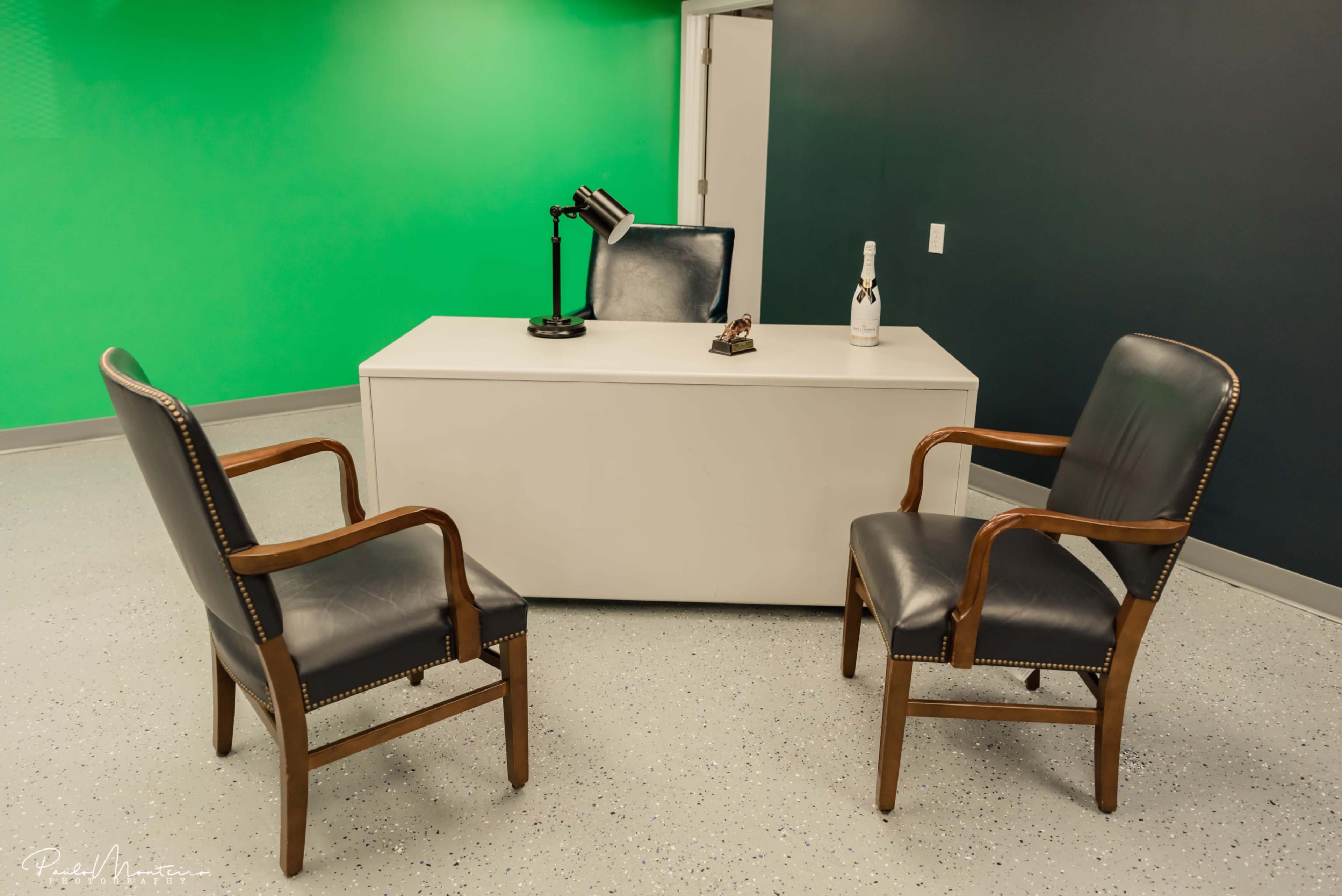 The image shows a small office space featuring a white desk, two wooden chairs, and a champagne bottle, with a green screen wall in the background.