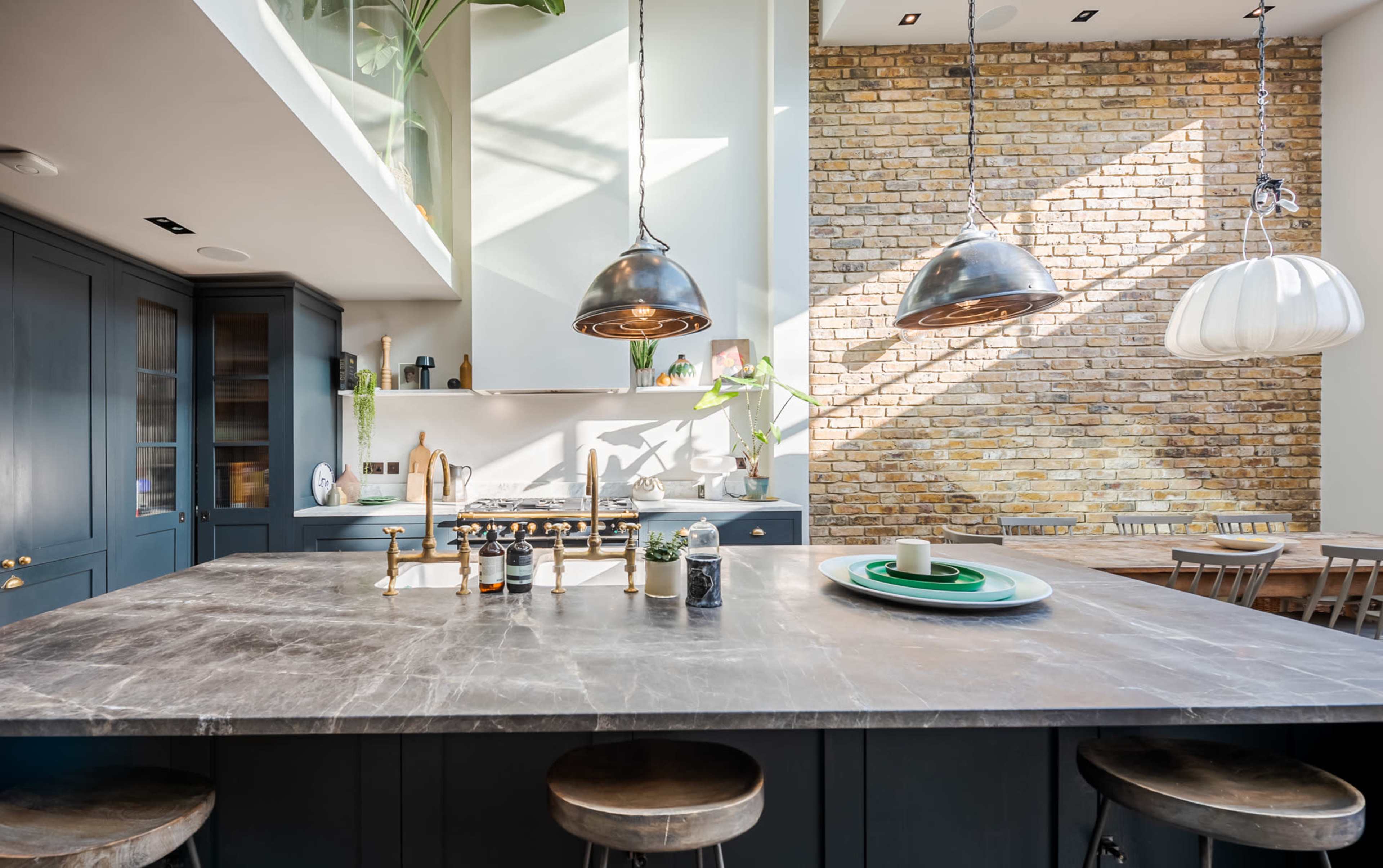 A modern kitchen features a large marble island with metal fixtures, pendant lighting, and a mix of cabinetry and exposed brick walls.