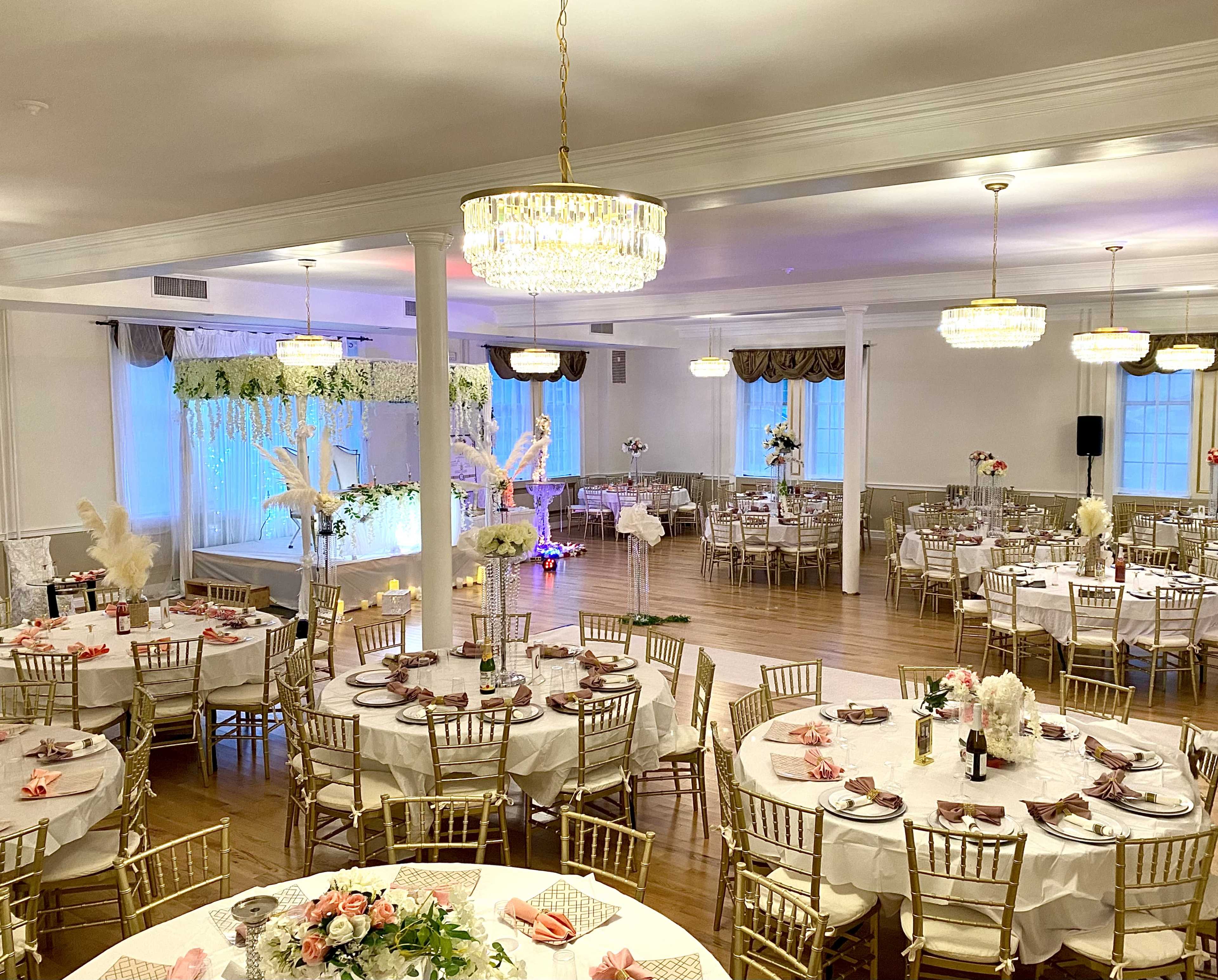 A decorated banquet hall with round tables set for an event, featuring elegant chairs, floral centerpieces, and soft, ambient lighting.