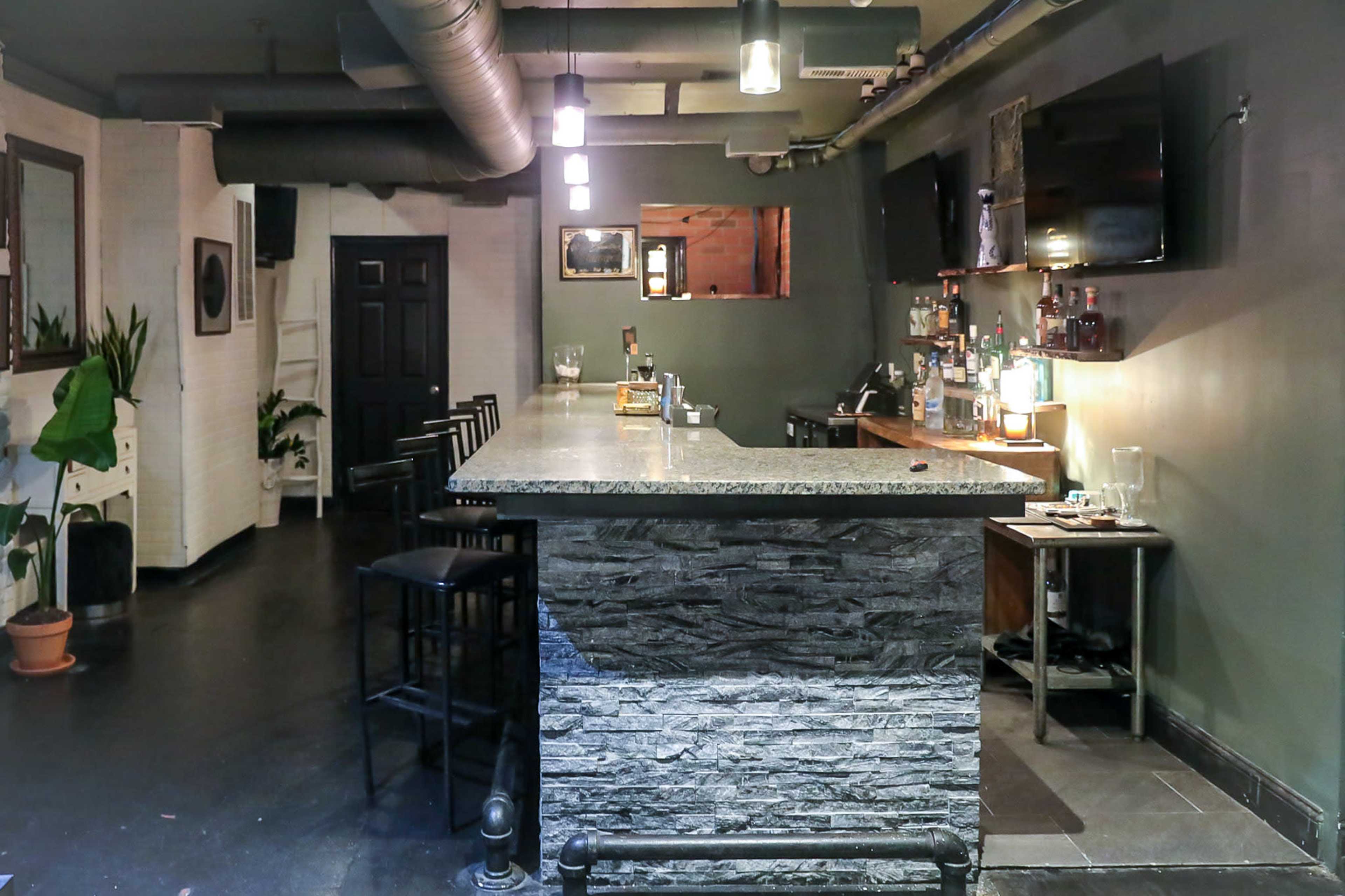 The image shows a dimly lit bar area with a granite countertop, high stools, and shelves displaying various bottles.
