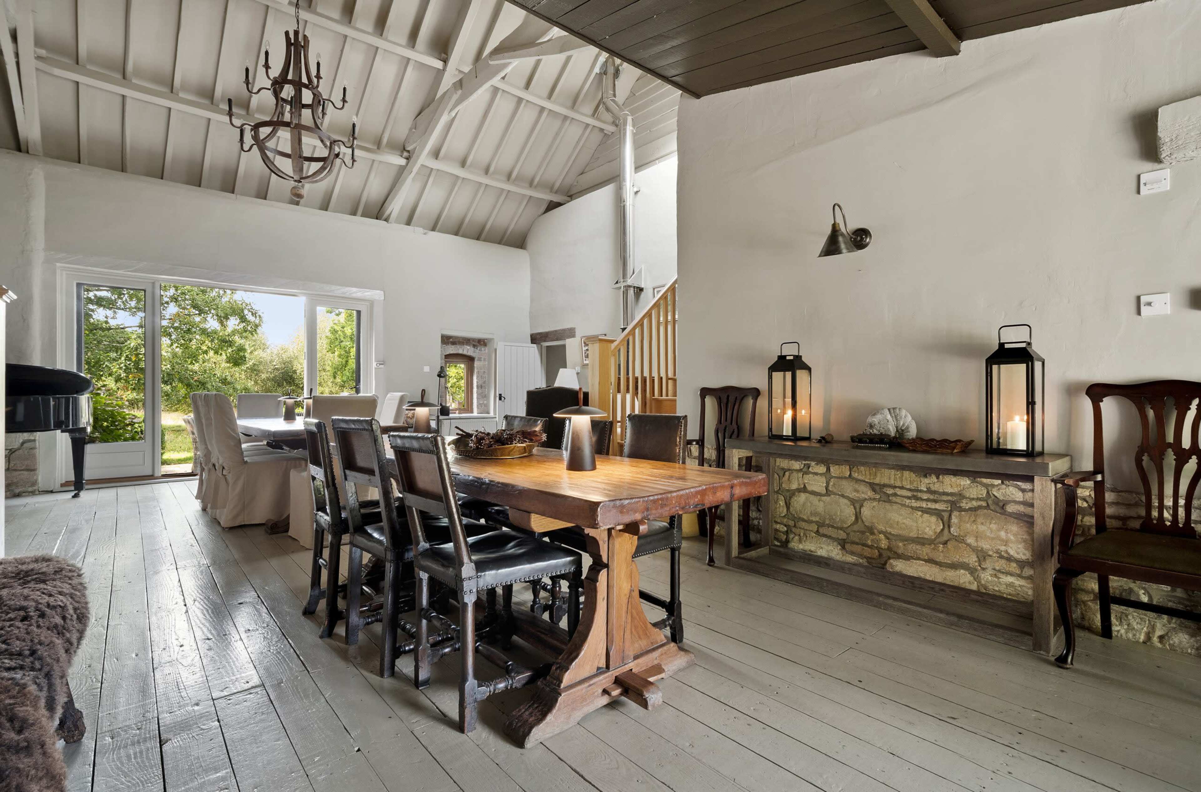 The image shows a spacious dining area with a wooden table and chairs, large windows overlooking greenery, and a stone-accented console table with lanterns.