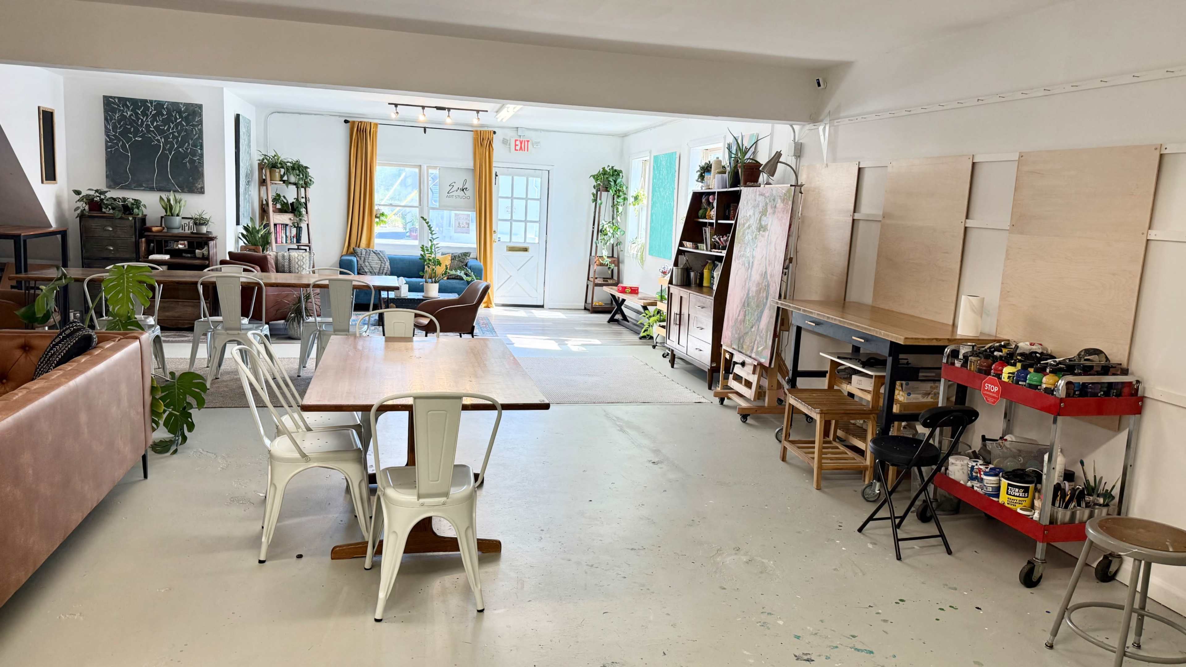The image shows a spacious, well-lit interior with a mix of furniture, including tables and chairs, and various plants, creating a vibrant workspace.