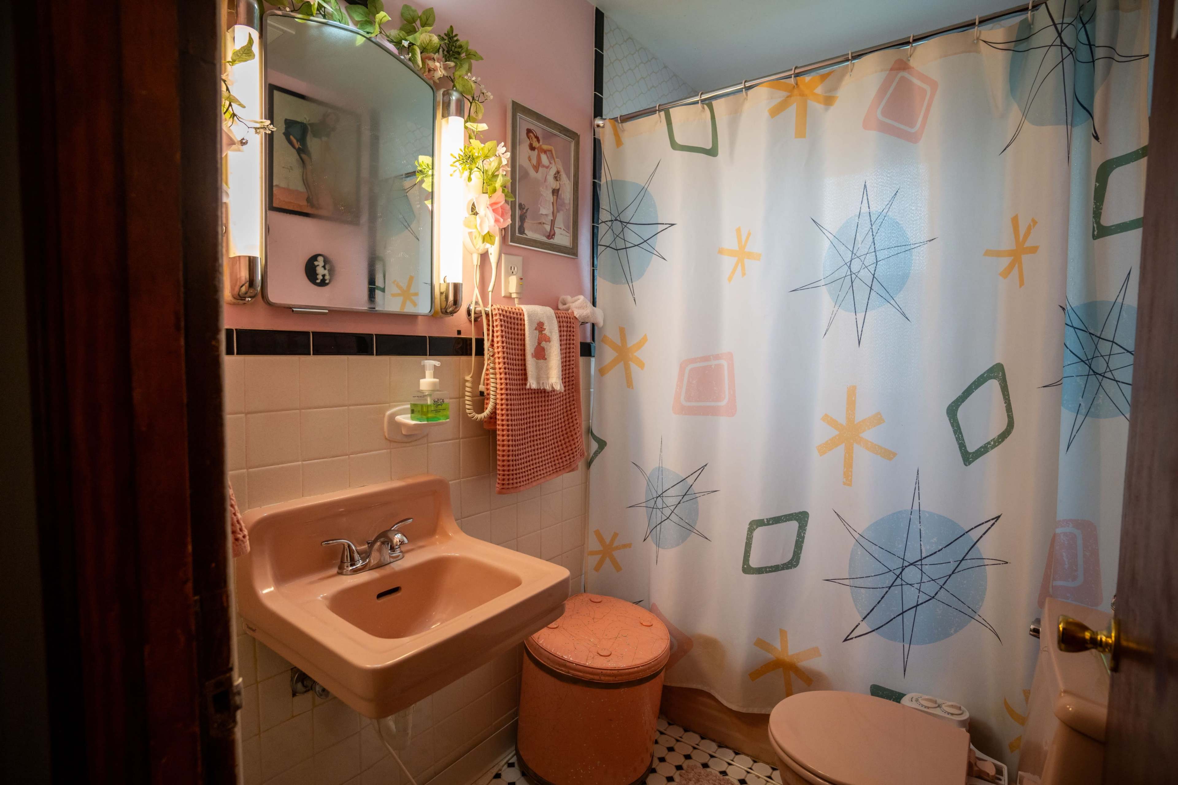 A small bathroom with a pink sink and toilet, a patterned shower curtain, and a mirror reflecting the room.