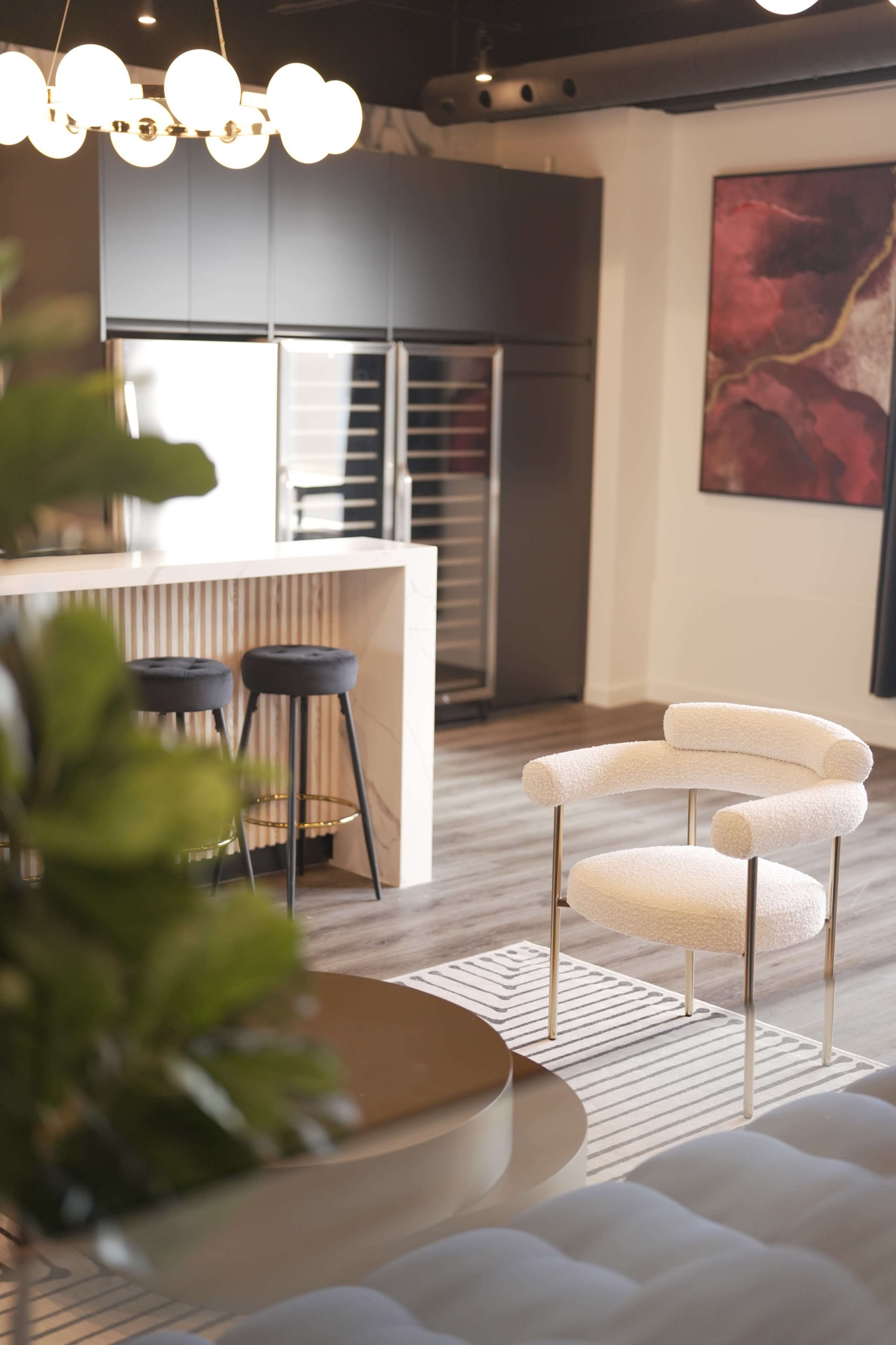 The image shows a modern kitchen and living area featuring a light-colored upholstered chair, a round table, and a dark cabinet with a wine refrigerator.