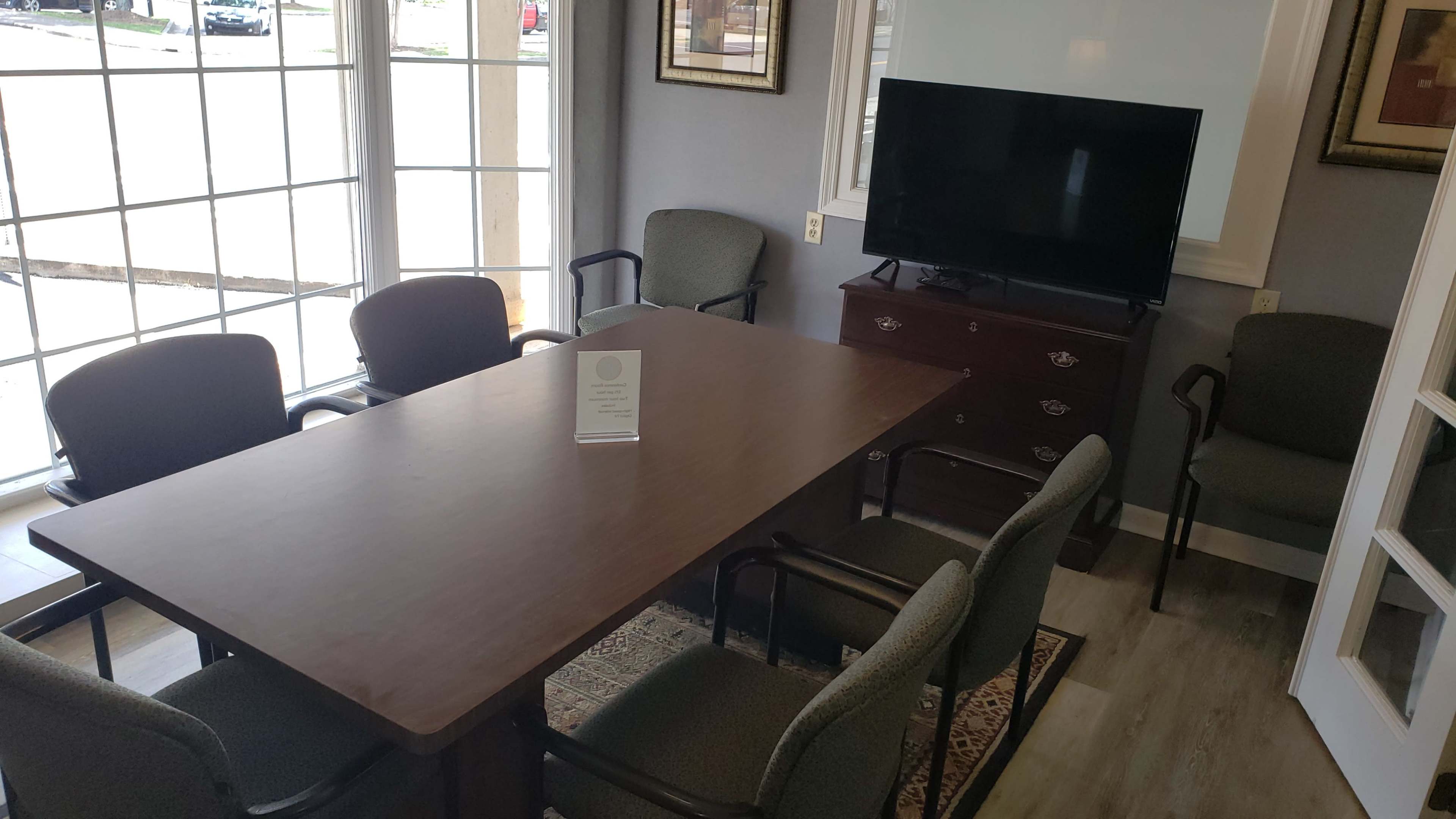 A conference room features a large wooden table surrounded by six chairs, with a television mounted on the wall and large windows allowing natural light.