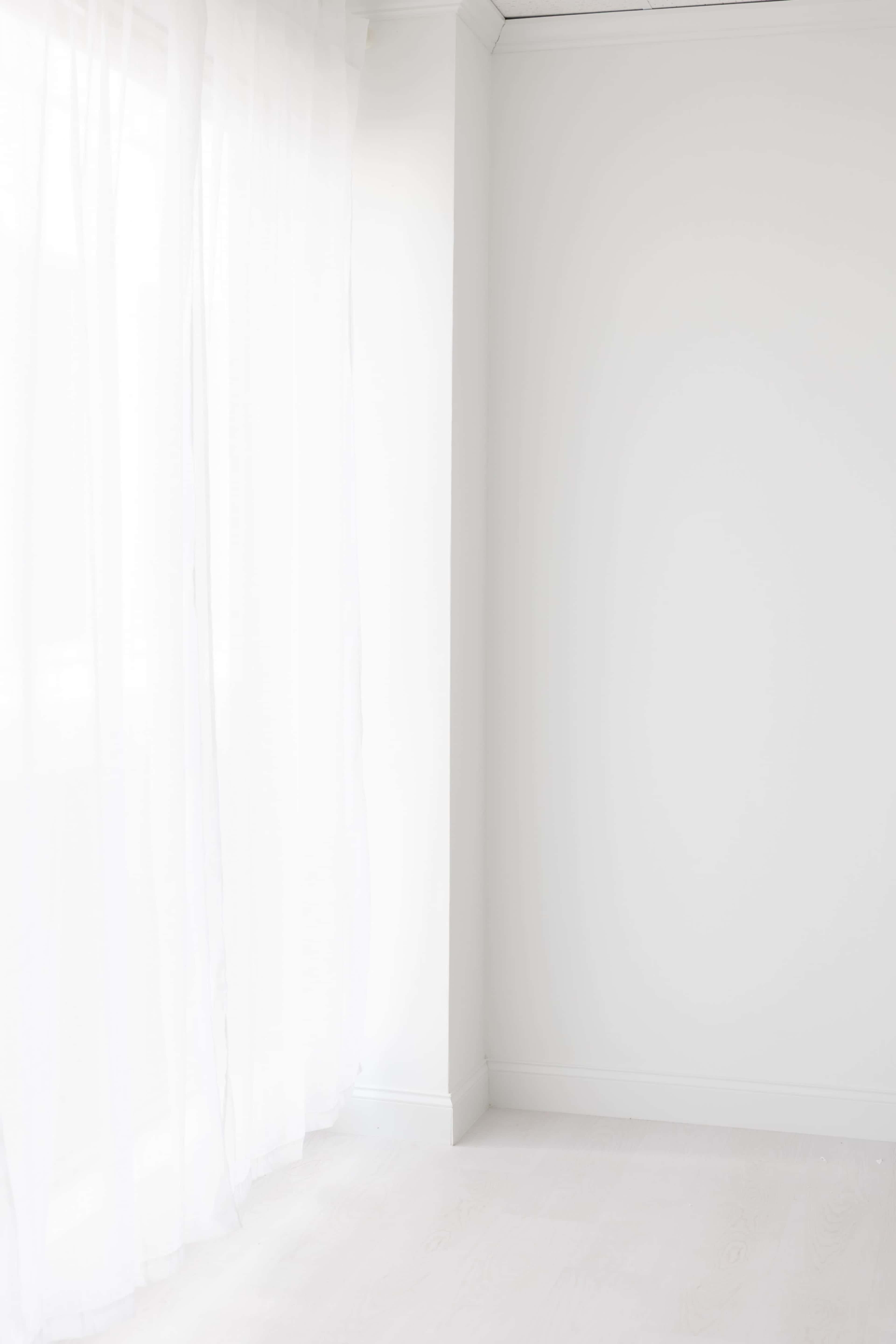 A bright room features a corner with white walls and sheer curtains letting in natural light.