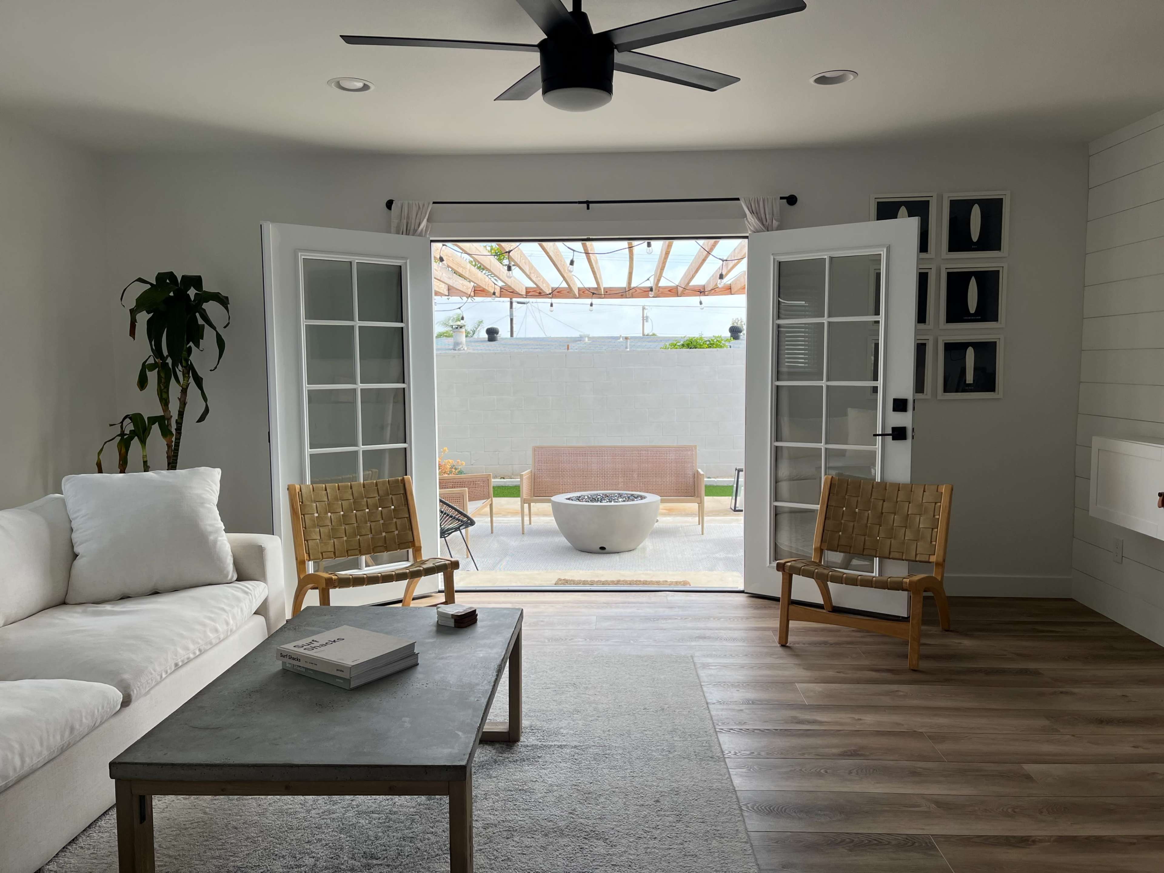 A bright living room features a white sofa, two wooden chairs, and French doors leading to a patio with a stone fire pit and outdoor seating.