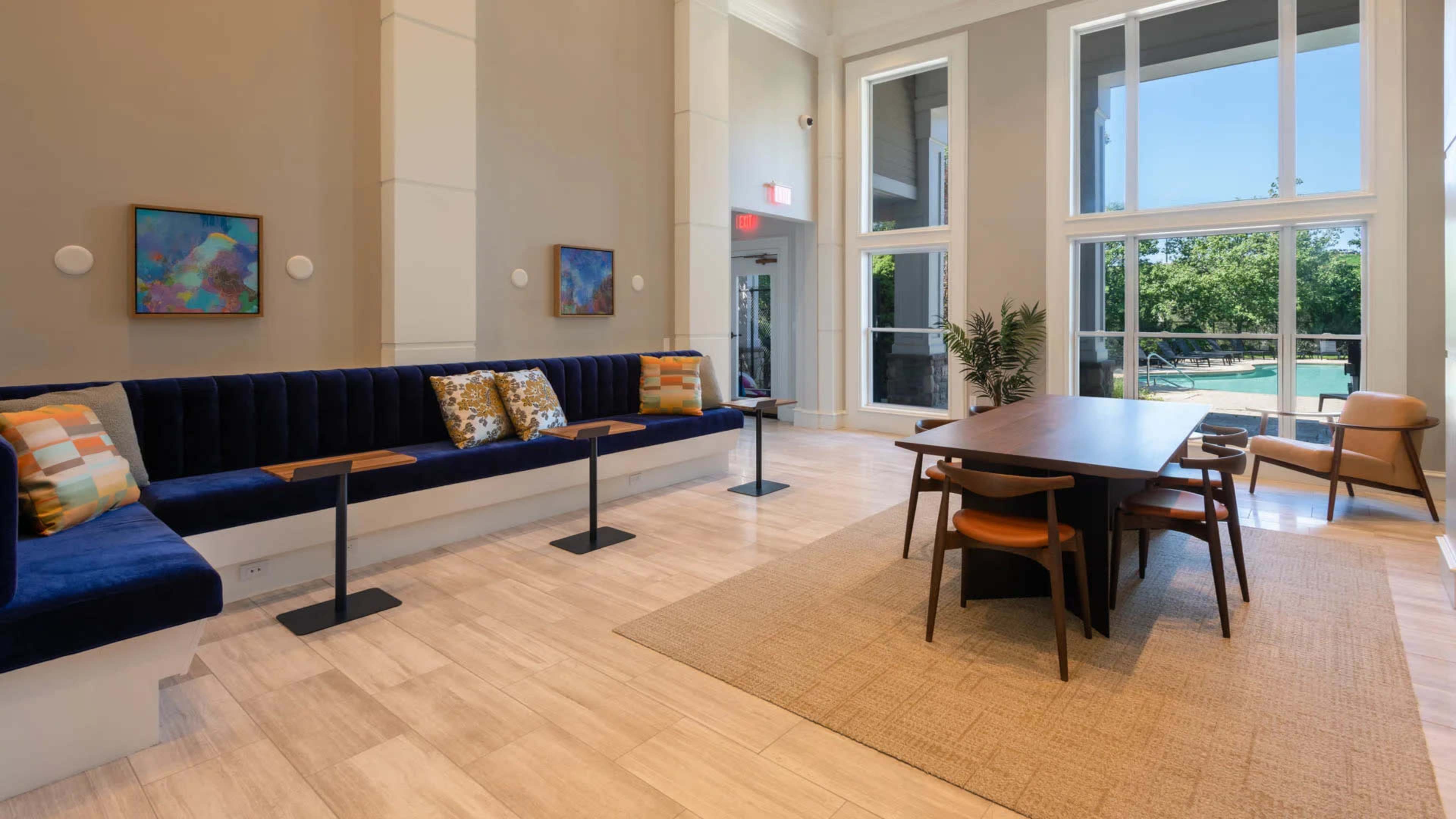 The image shows a modern lounge area featuring a blue upholstered bench, a wooden dining table with chairs, and large windows that overlook greenery.