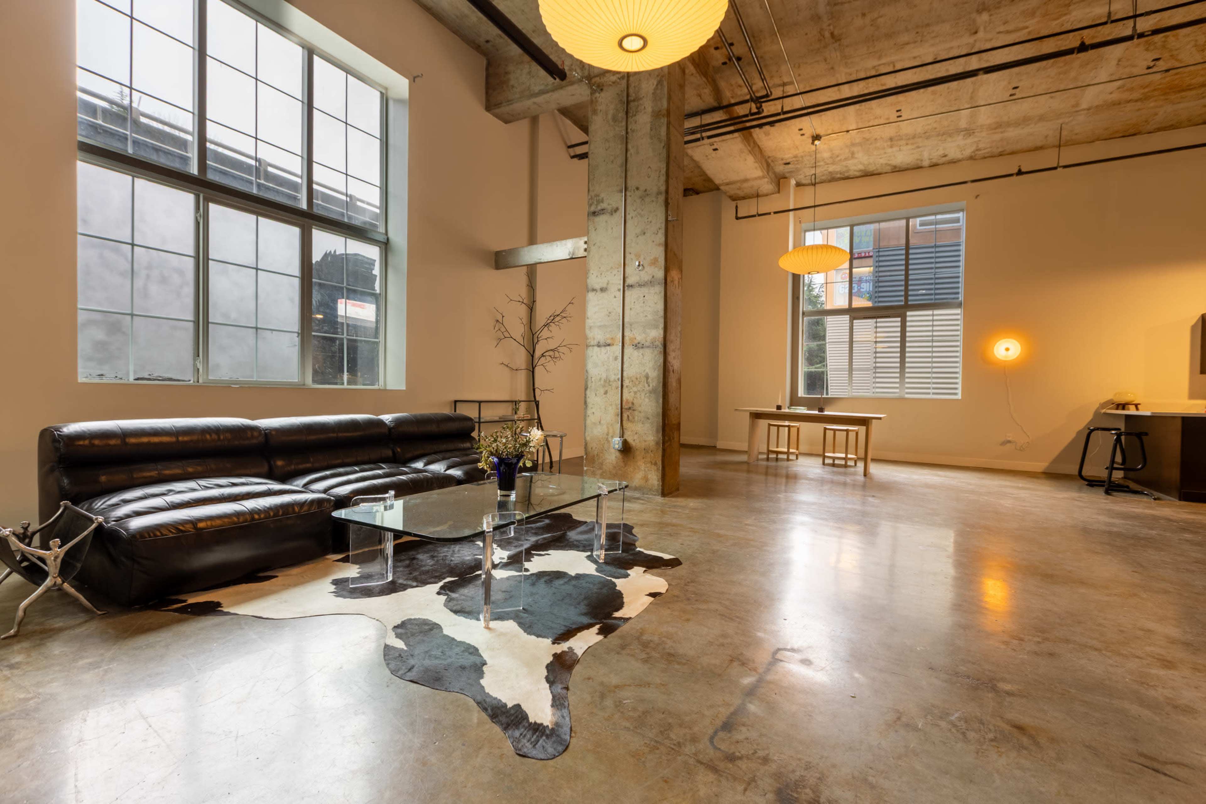 The image shows a spacious, modern living room with a concrete floor, large windows, and minimalist furniture, including a black leather sofa and a glass coffee table.