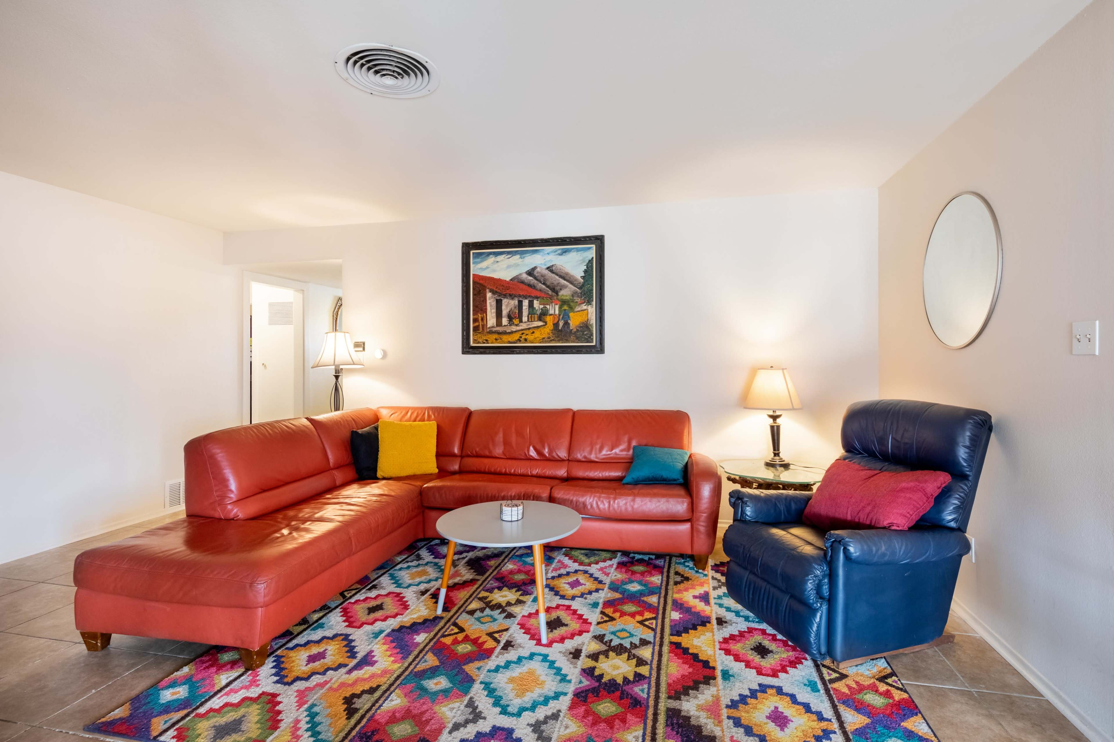 The room features a vibrant red sectional sofa, a blue armchair, a round coffee table, and a colorful patterned rug, with a painting on the wall and lamps providing light.