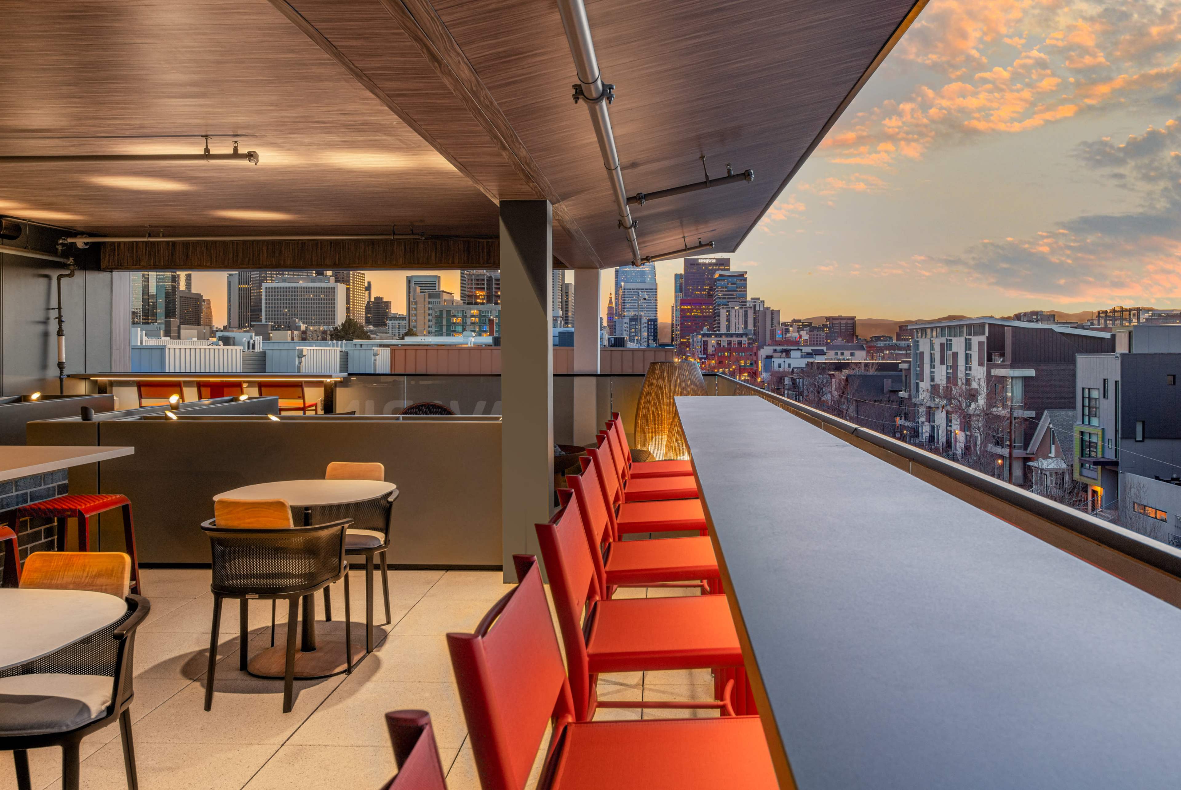 A modern rooftop bar with red chairs and a long gray countertop, overlooking a city skyline at sunset.
