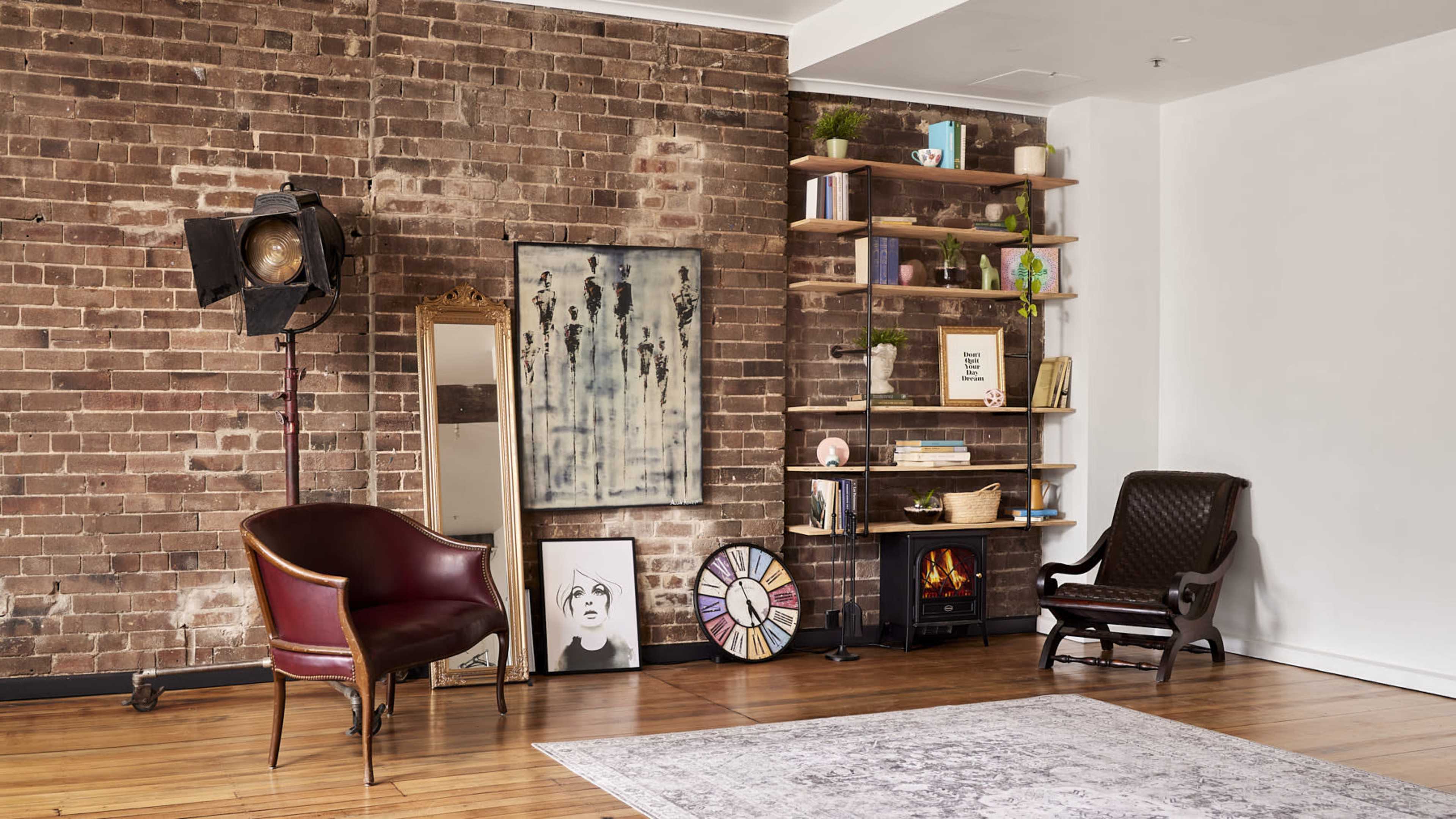 A stylish interior with exposed brick walls, a vintage armchair, a wooden bookshelf filled with decorative items, and a small electric fireplace.