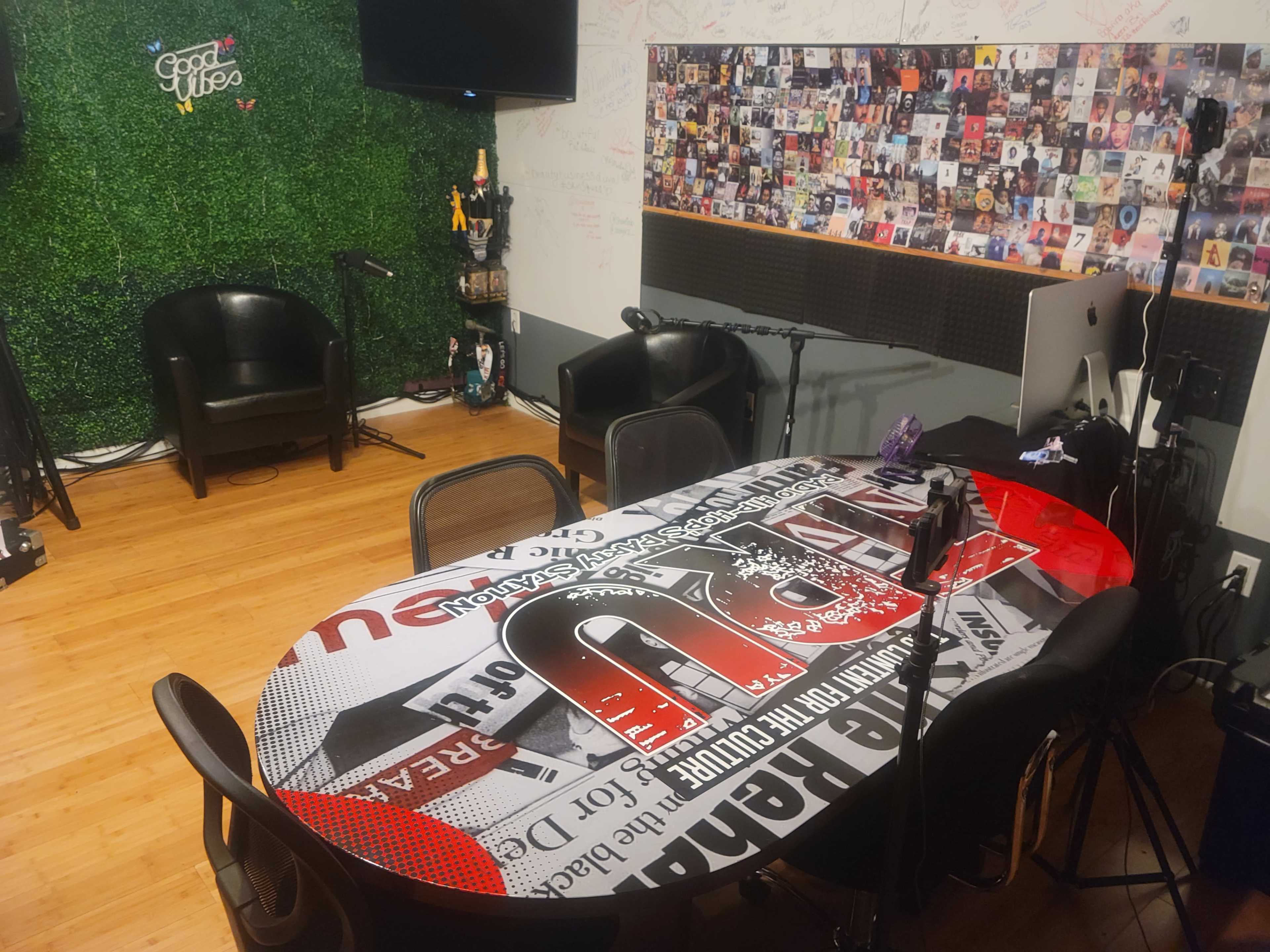 A media recording studio with a round table covered in graphic designs, black chairs, and walls adorned with framed photographs and green foliage.