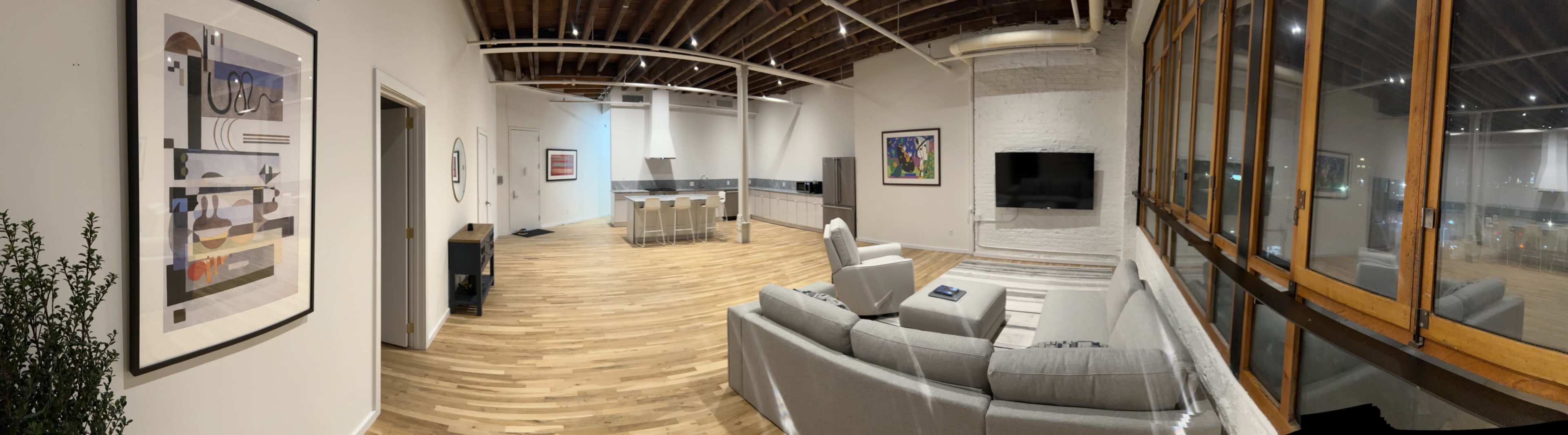 The image shows a spacious, modern interior with a living area featuring a gray sofa, a television, and a kitchen with an island, all surrounded by large windows.