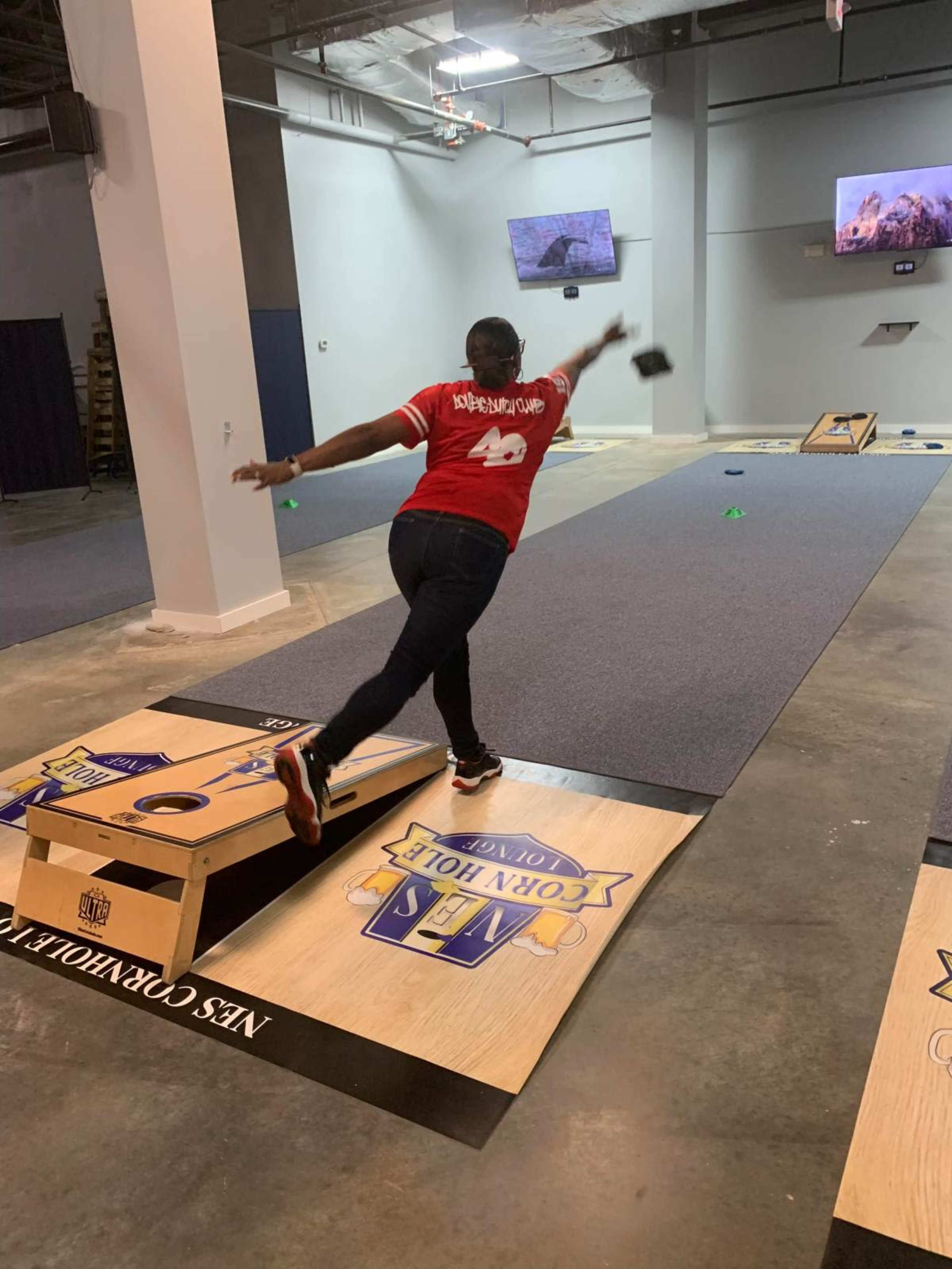 A person in a red shirt throws a bean bag onto a cornhole board in a recreational space.