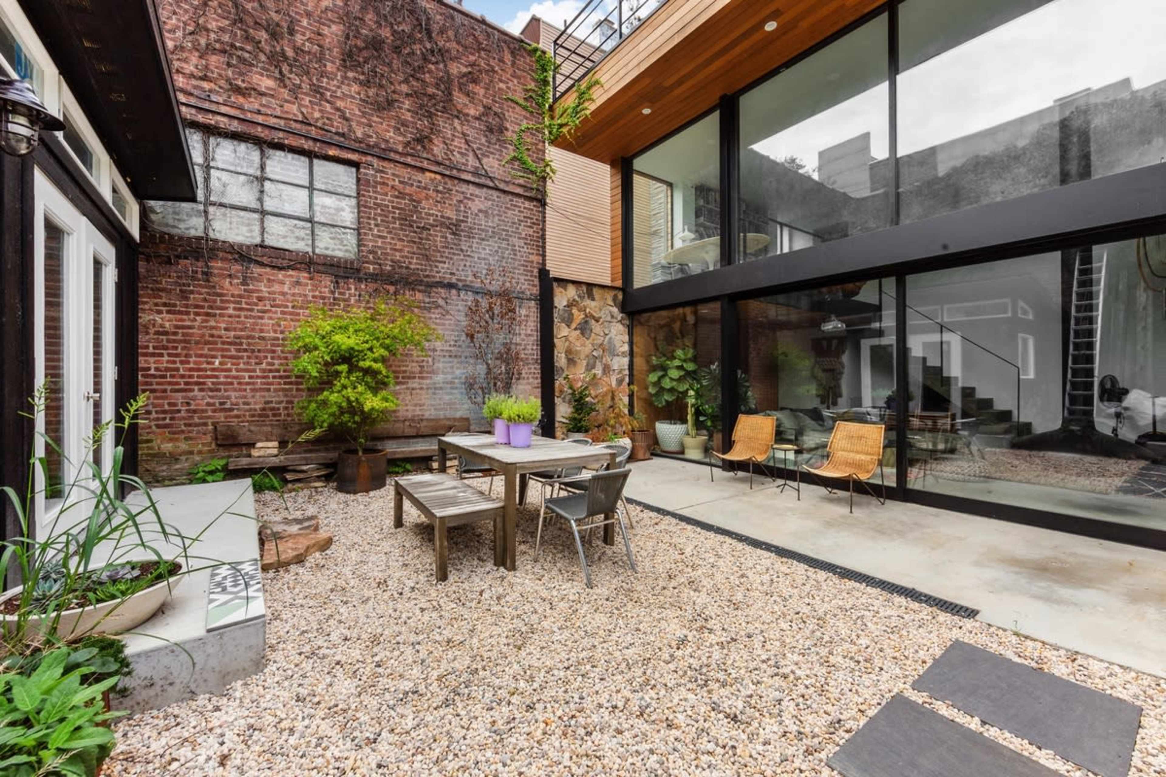 The image features a modern courtyard with a gravel floor, surrounded by a mix of brick and glass walls, and includes a wooden table, metal chairs, and potted plants.