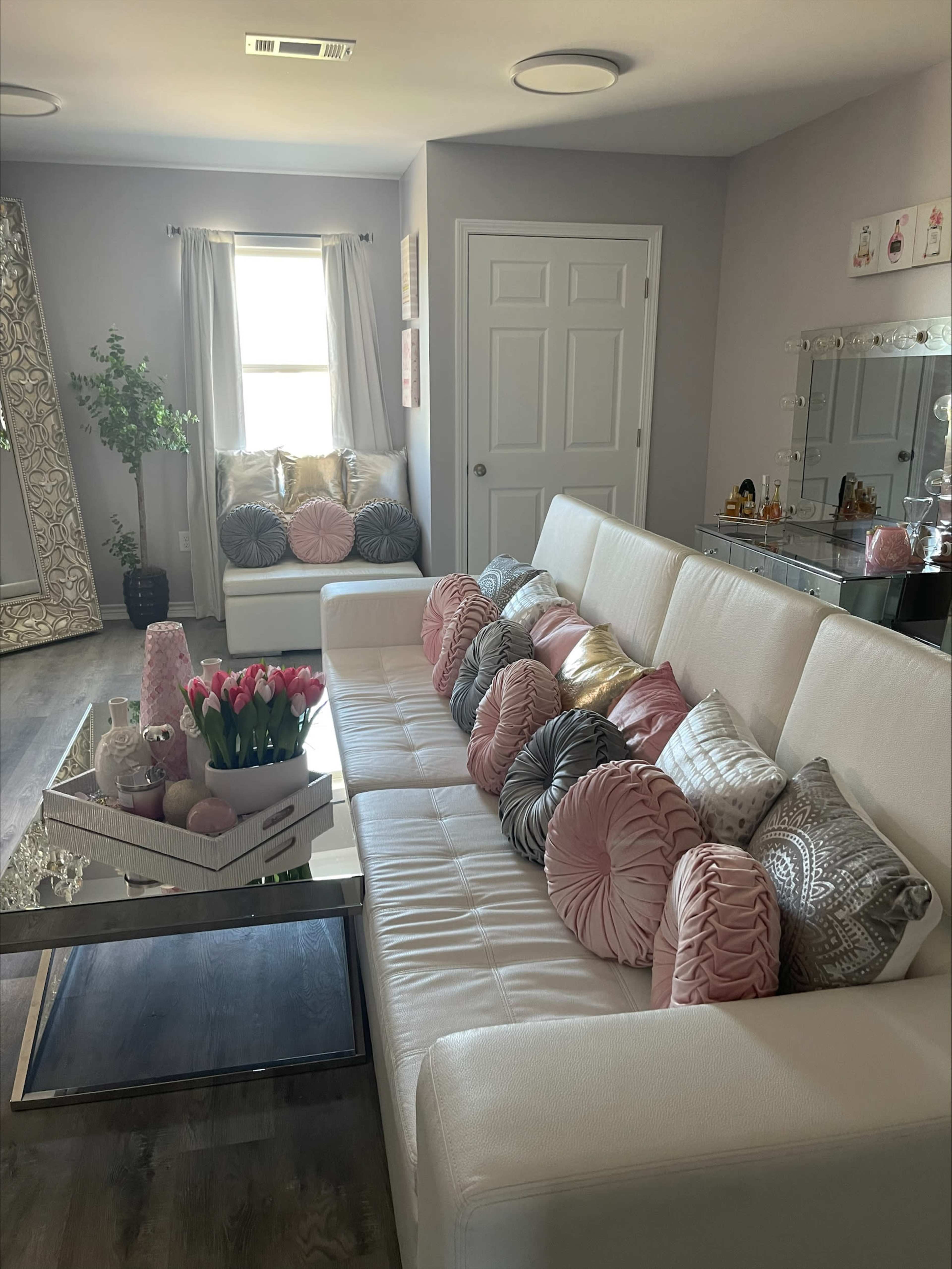 A modern living room featuring a white sofa adorned with various decorative pillows, a glass coffee table with a floral centerpiece, and a mirror with a decorative frame.
