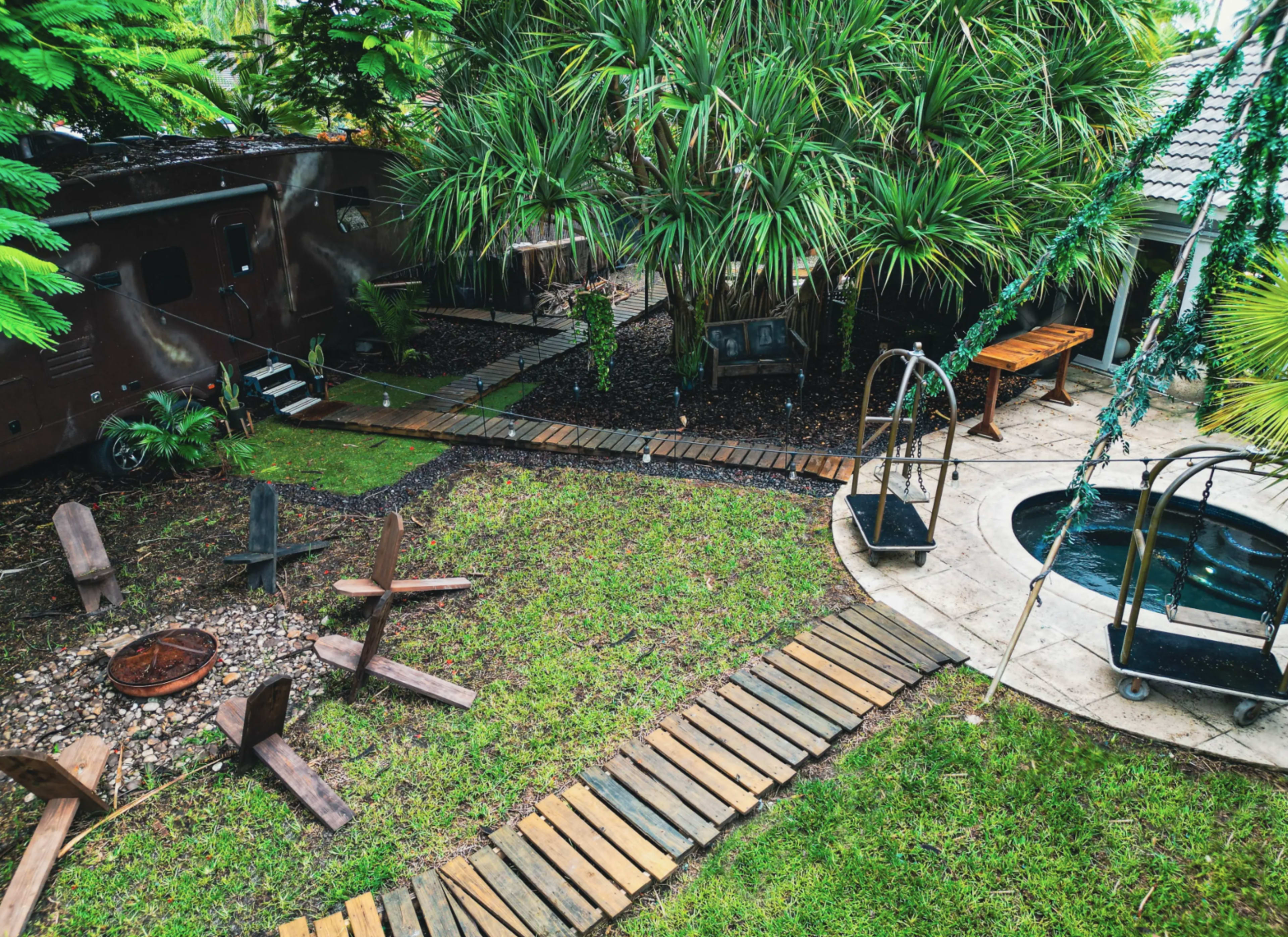 The image shows a landscaped yard featuring wooden paths, a fire pit surrounded by seating, a hot tub, and lush greenery.