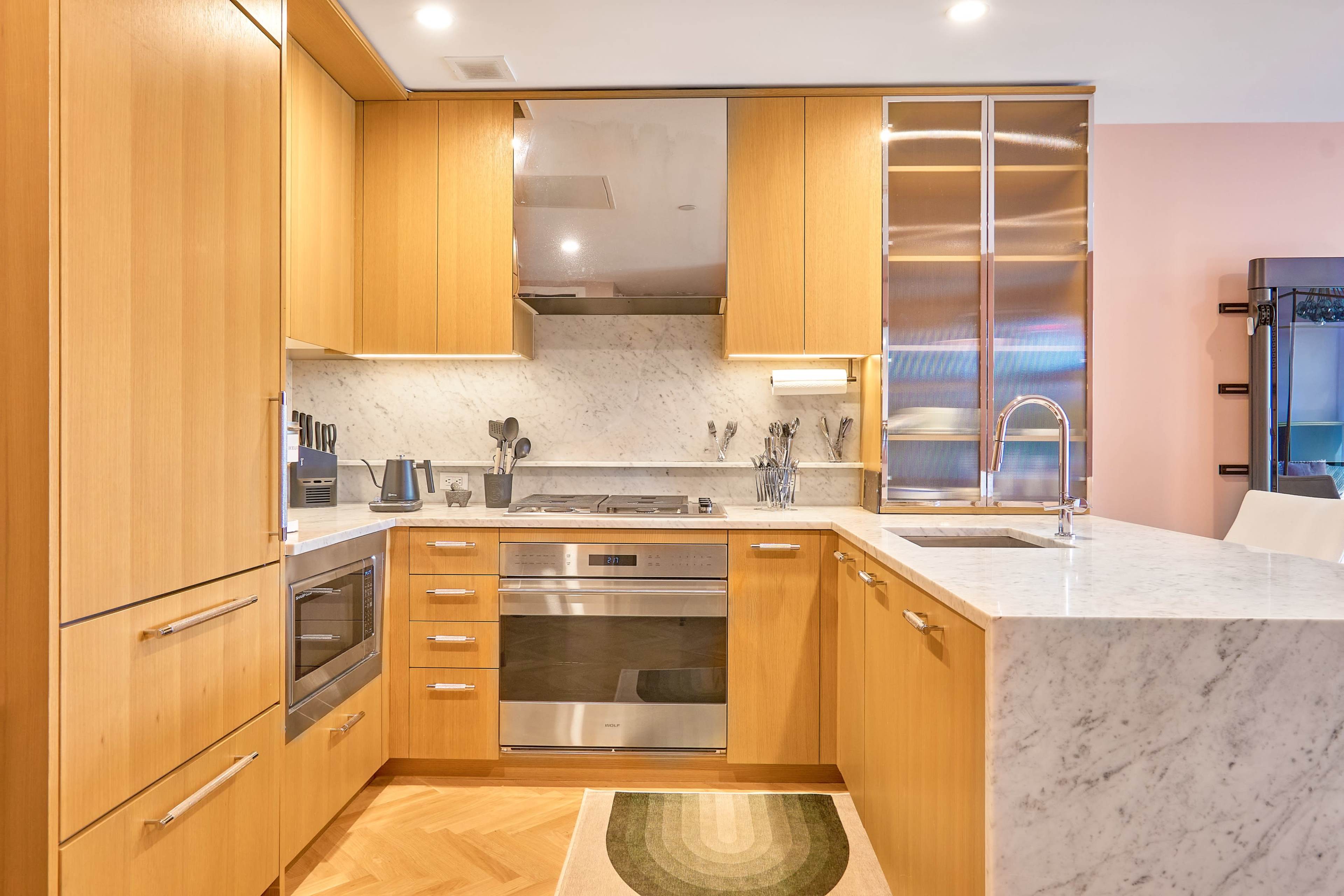 A modern kitchen features wooden cabinetry, stainless steel appliances, and a marble countertop with a patterned rug on the floor.