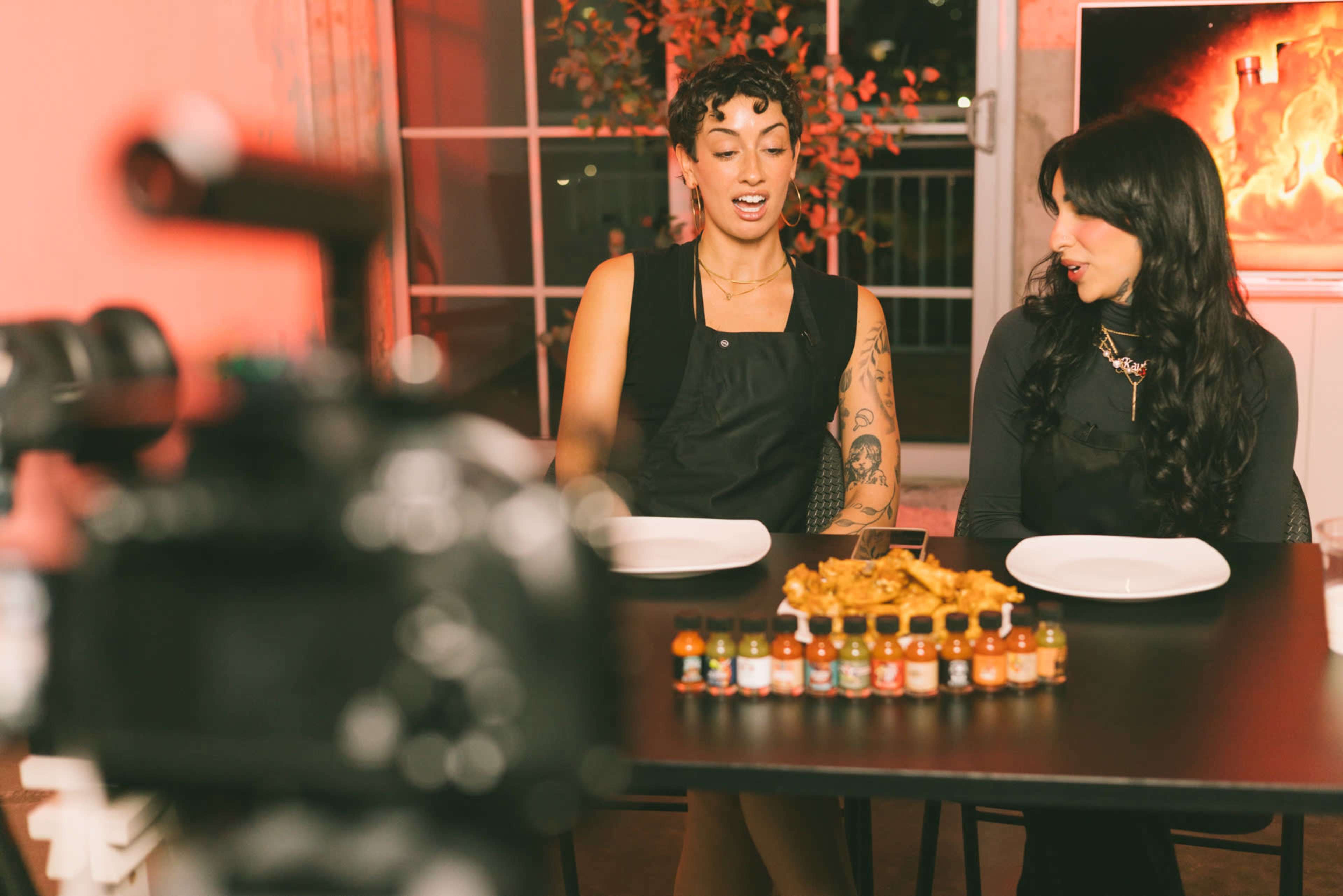 Two women are seated at a table with empty plates and a row of colorful hot sauce bottles in front of them, while a camera is positioned nearby.