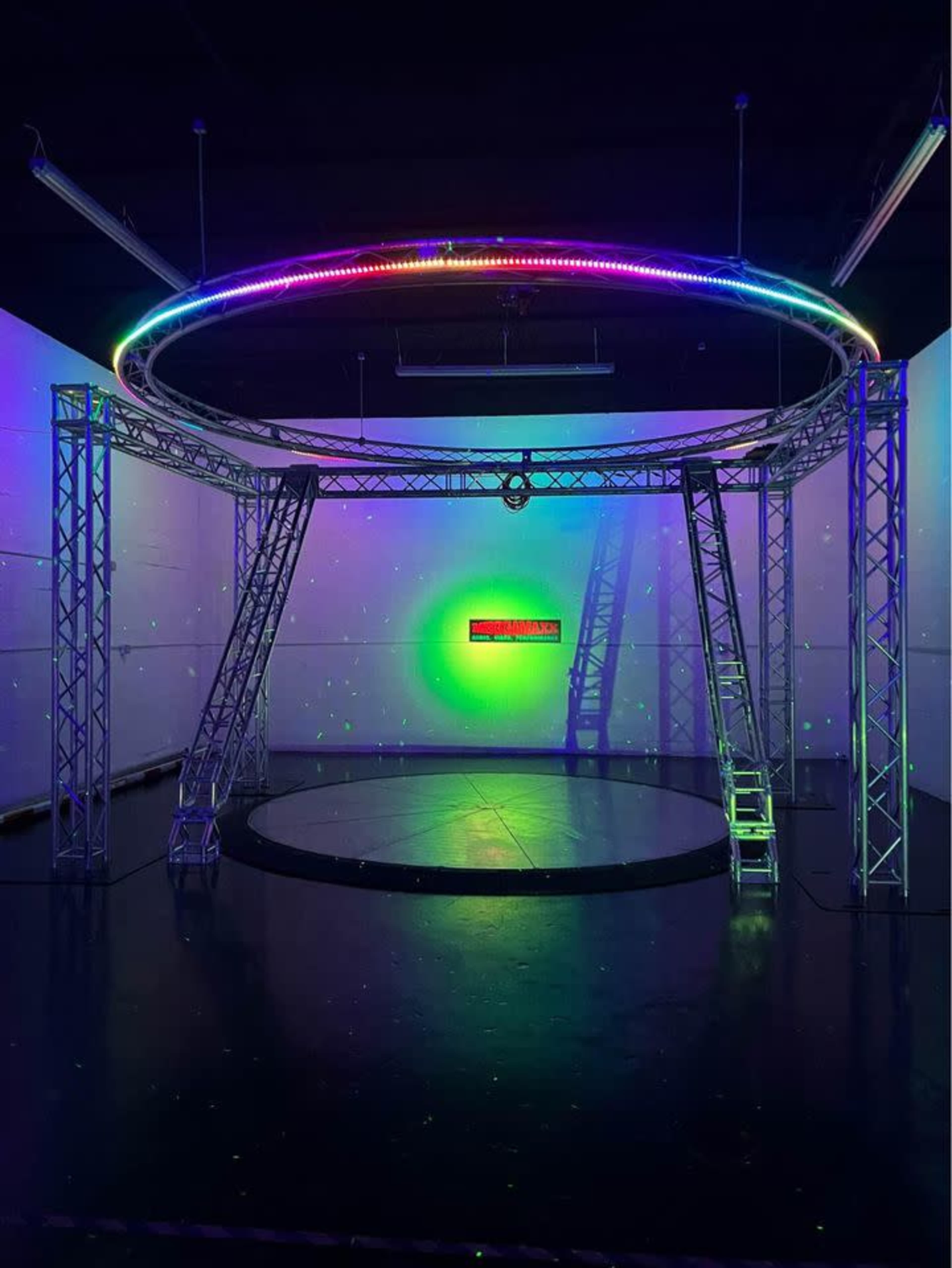 A circular stage structure with bright, multicolored lights and a dark floor in a dimly lit space.