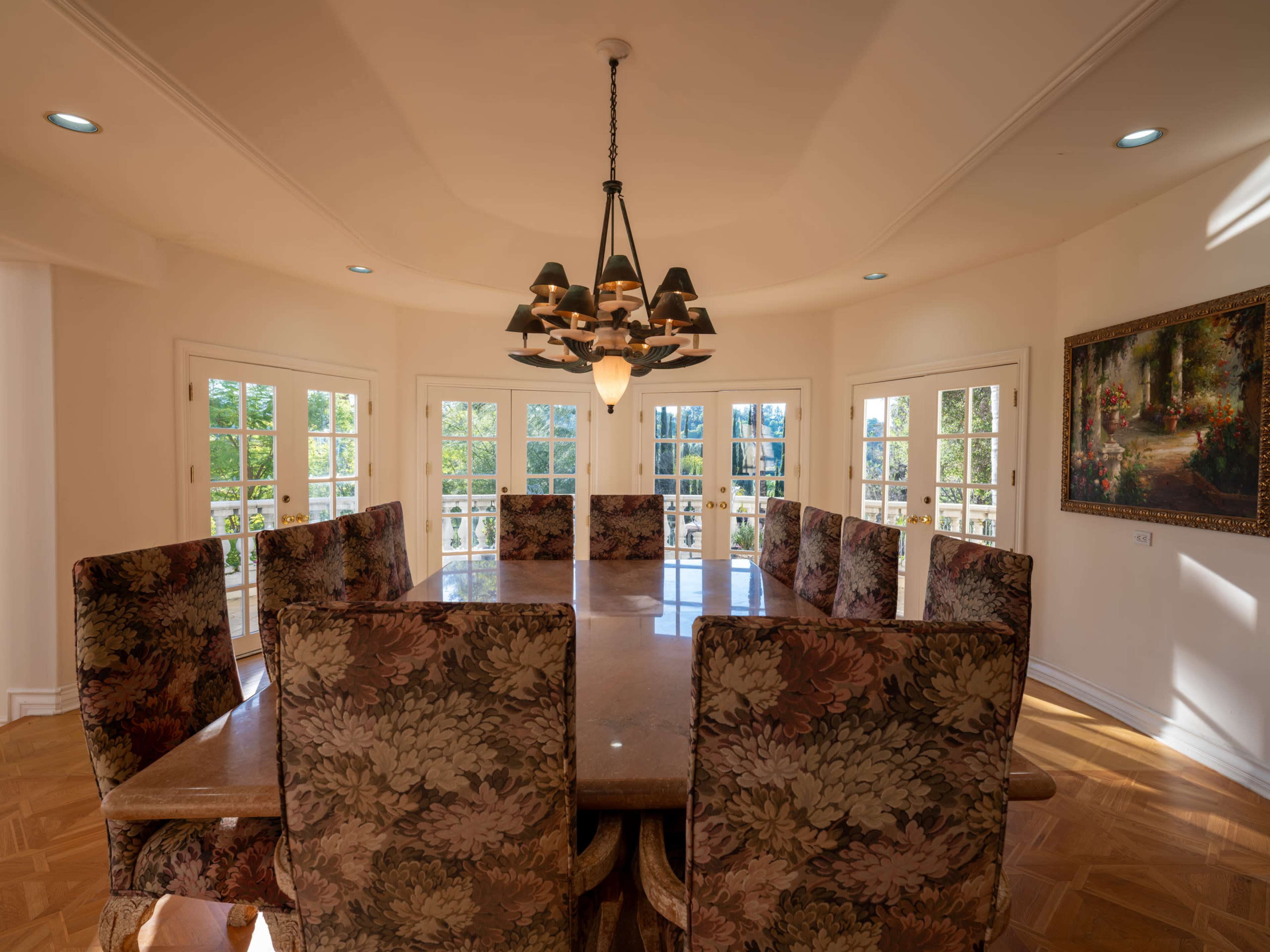 The image shows a spacious dining room with a large wooden table surrounded by upholstered floral chairs, illuminated by a central chandelier and featuring multiple windows providing natural light.