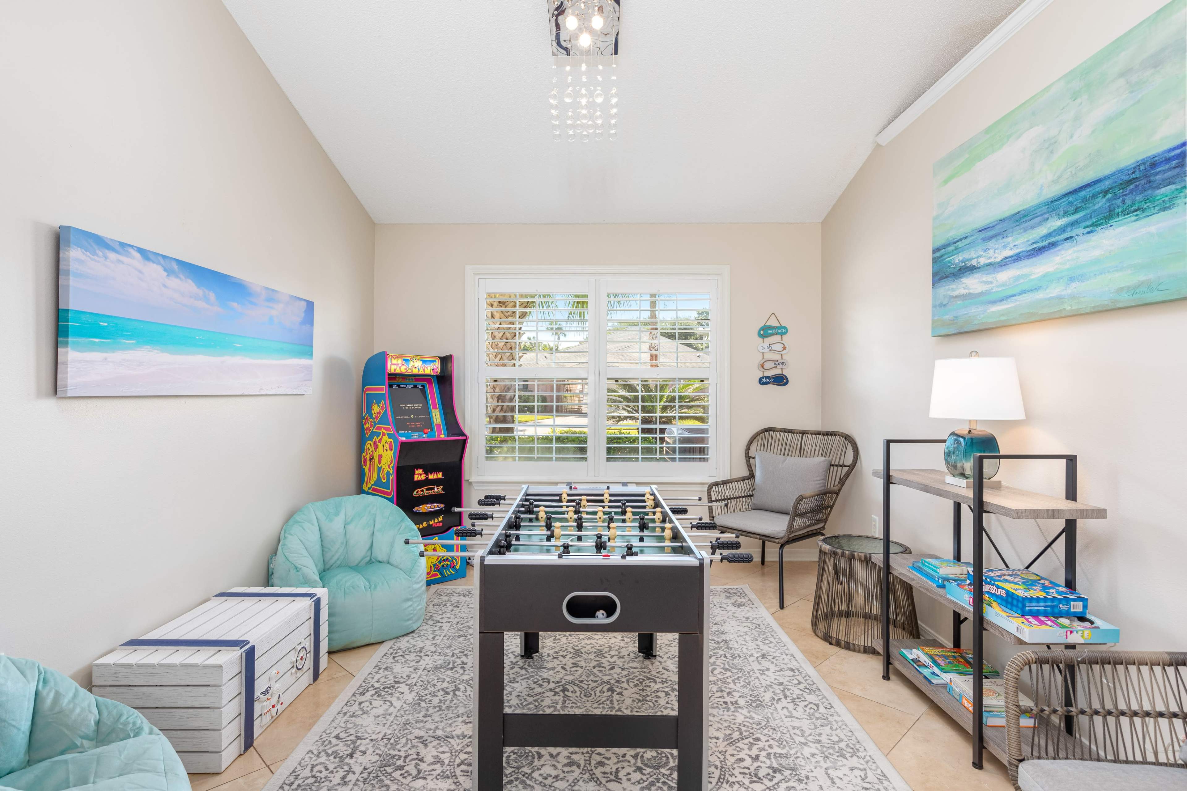 The room features a foosball table, arcade game, bean bag chairs, and a decorative lamp, with large windows allowing natural light to enter.