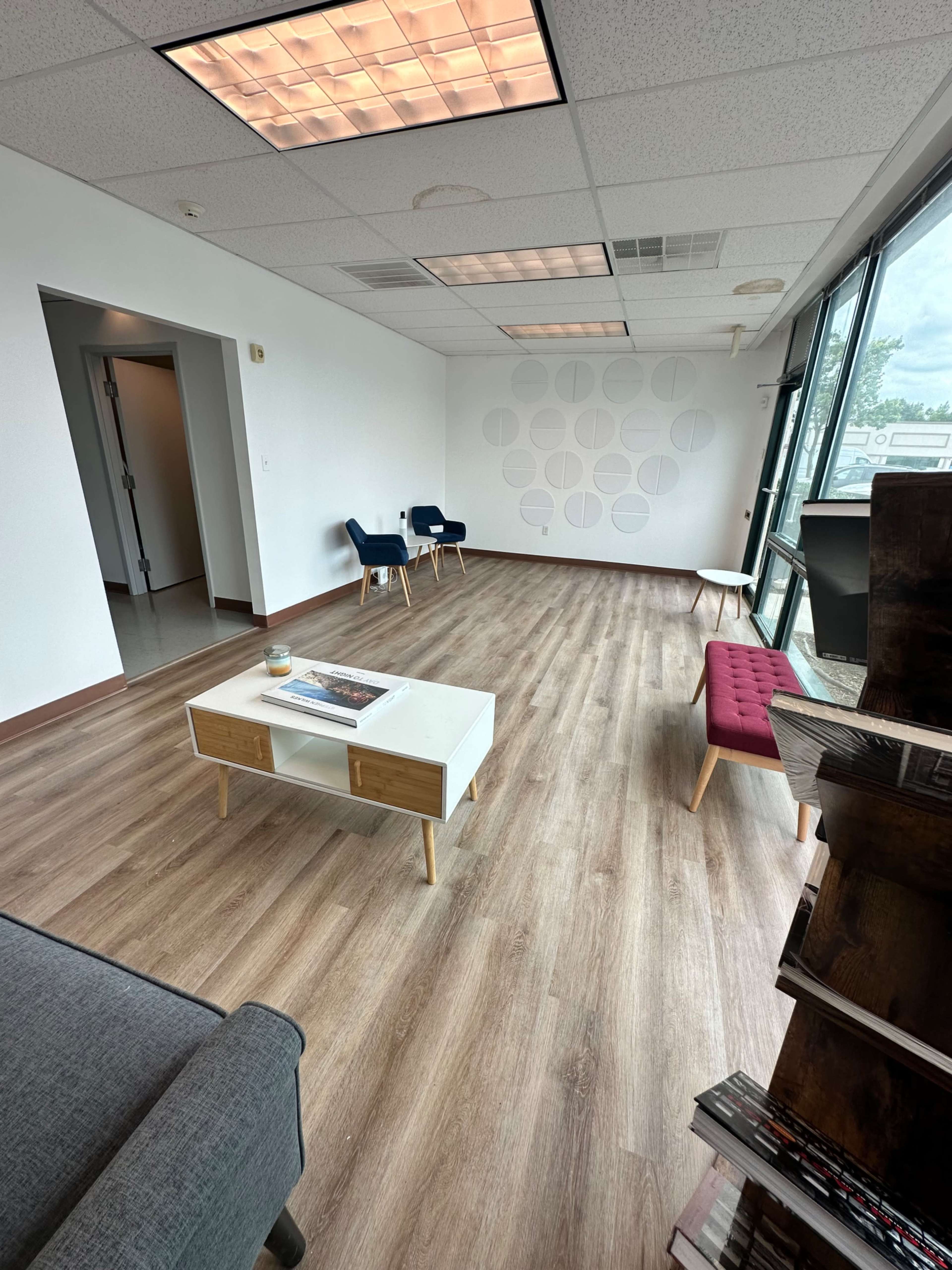 An empty office space with wooden flooring, a small coffee table, two chairs, and large windows.