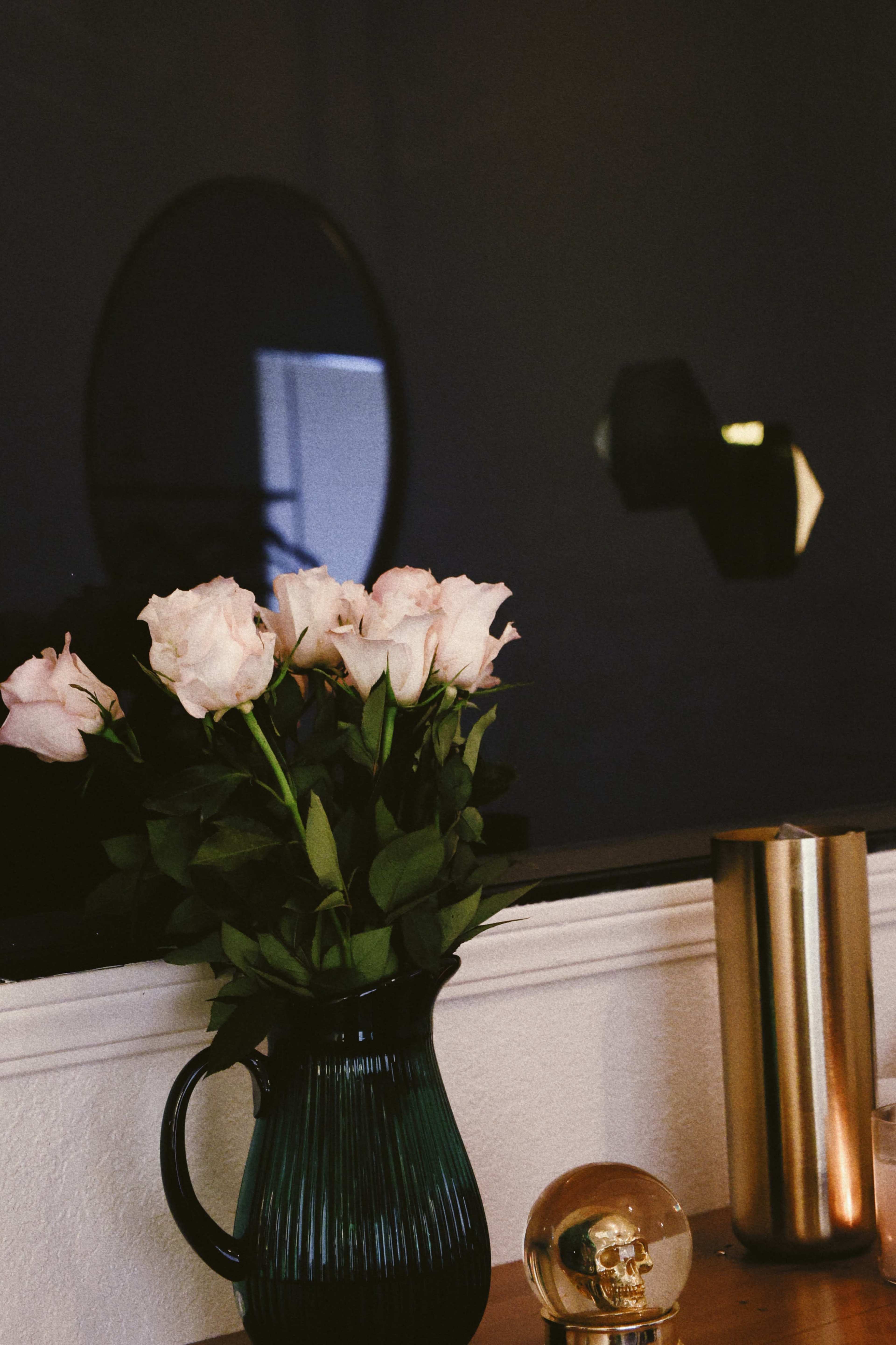 A vase of pink roses sits on a wooden table next to a decorative glass orb and a metal lamp, with a circular mirror reflecting light in the background.