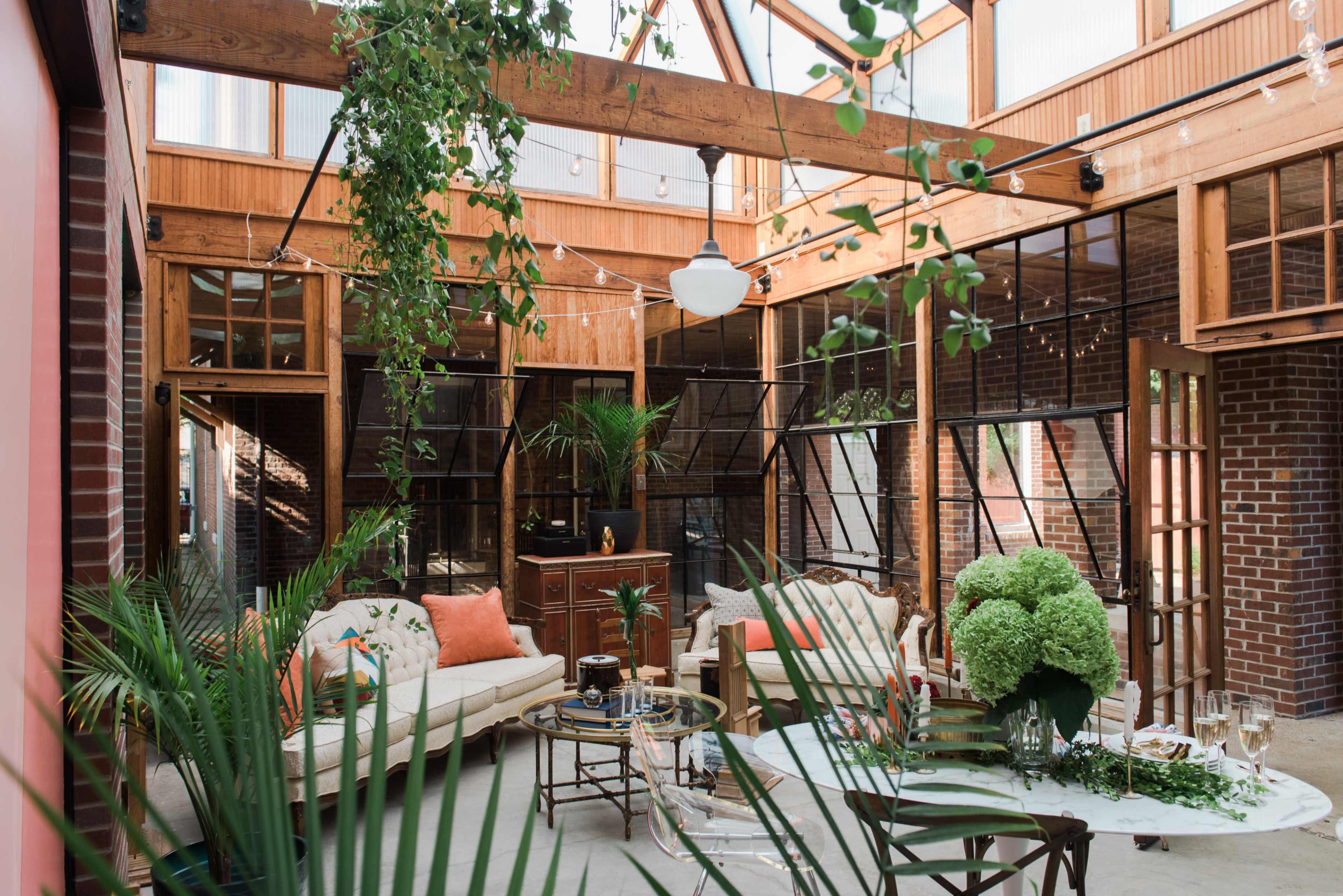 The image shows a bright and open indoor space featuring a living area with a sofa, plants, and a round dining table under a glass roof.