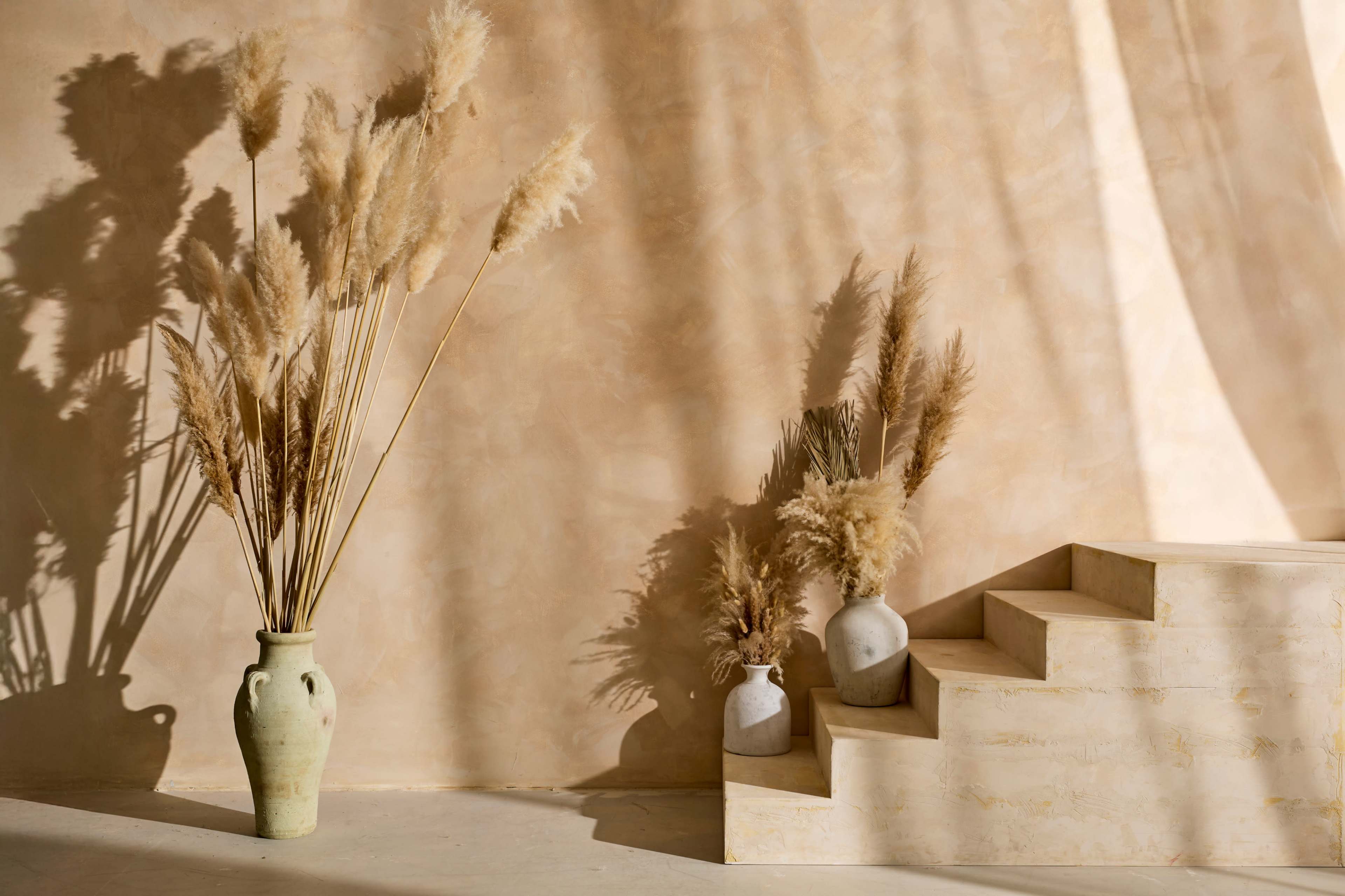 A vase filled with pampas grass stands beside a set of steps against a textured, warm-toned wall.