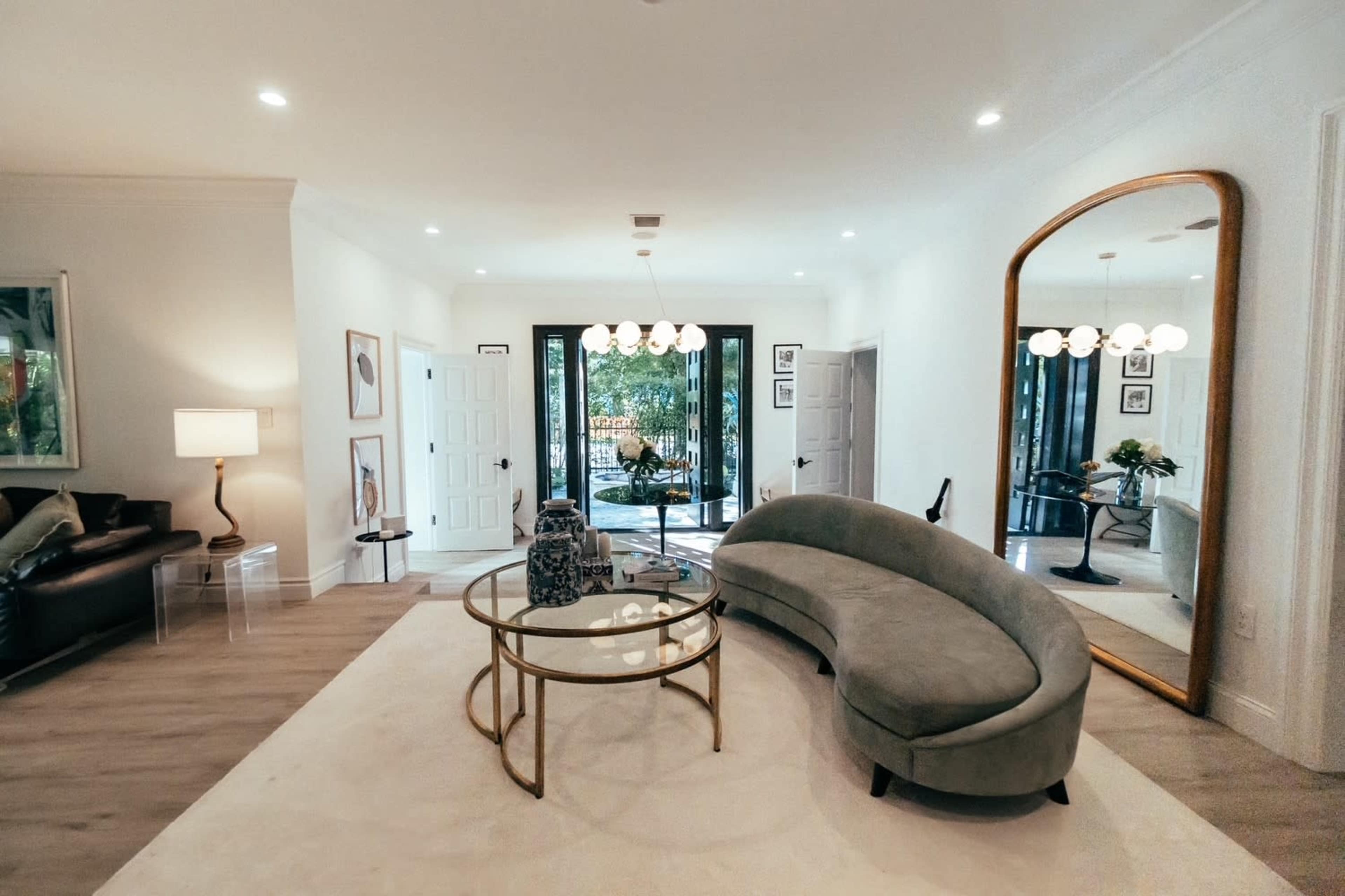 The image shows a modern living room with a curved gray sofa, a glass coffee table, and large doors leading to an outdoor space.