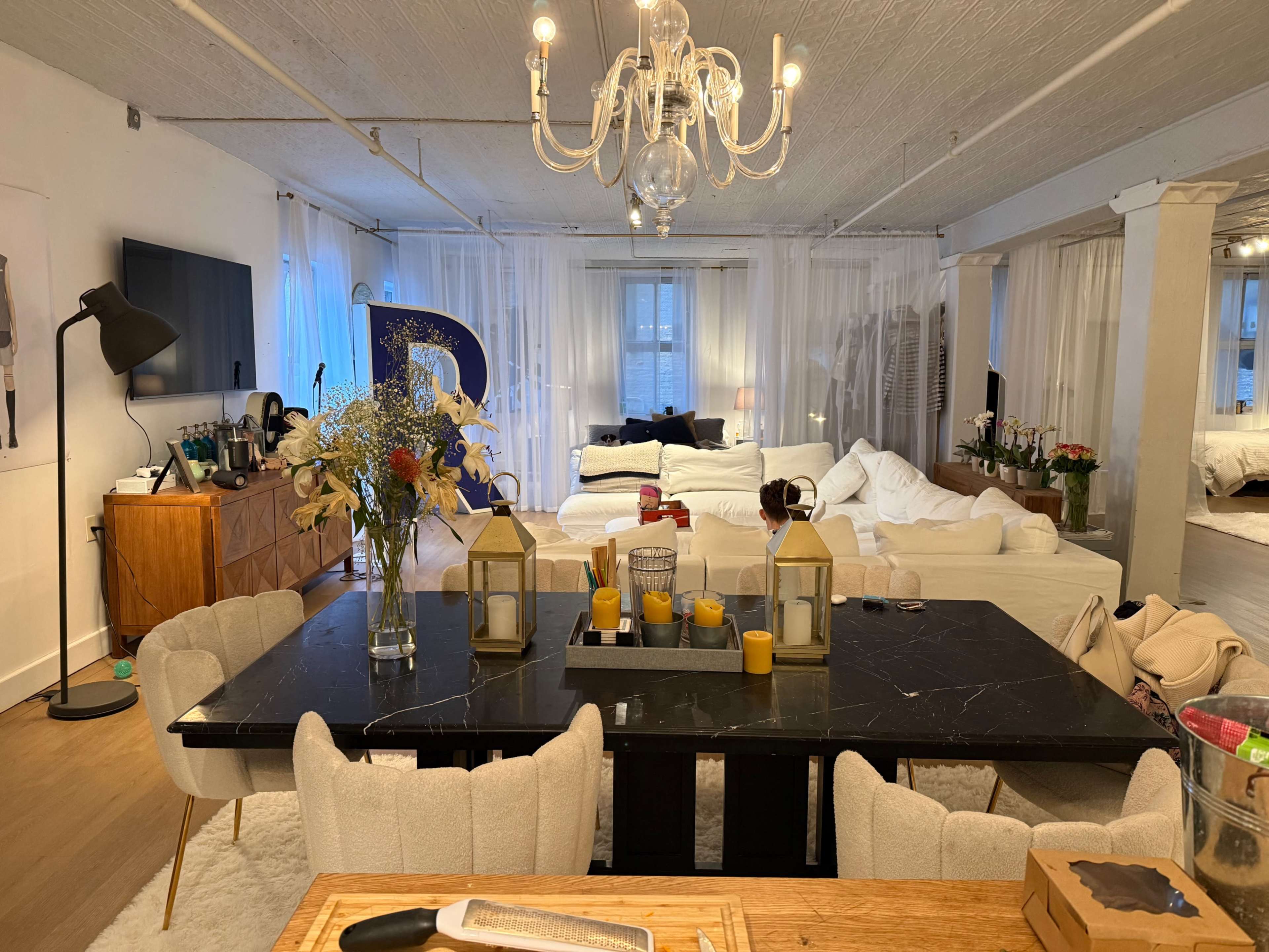 The image shows a spacious living area with a large black dining table, white sofas, a chandelier, and decorative elements like flowers and candles.