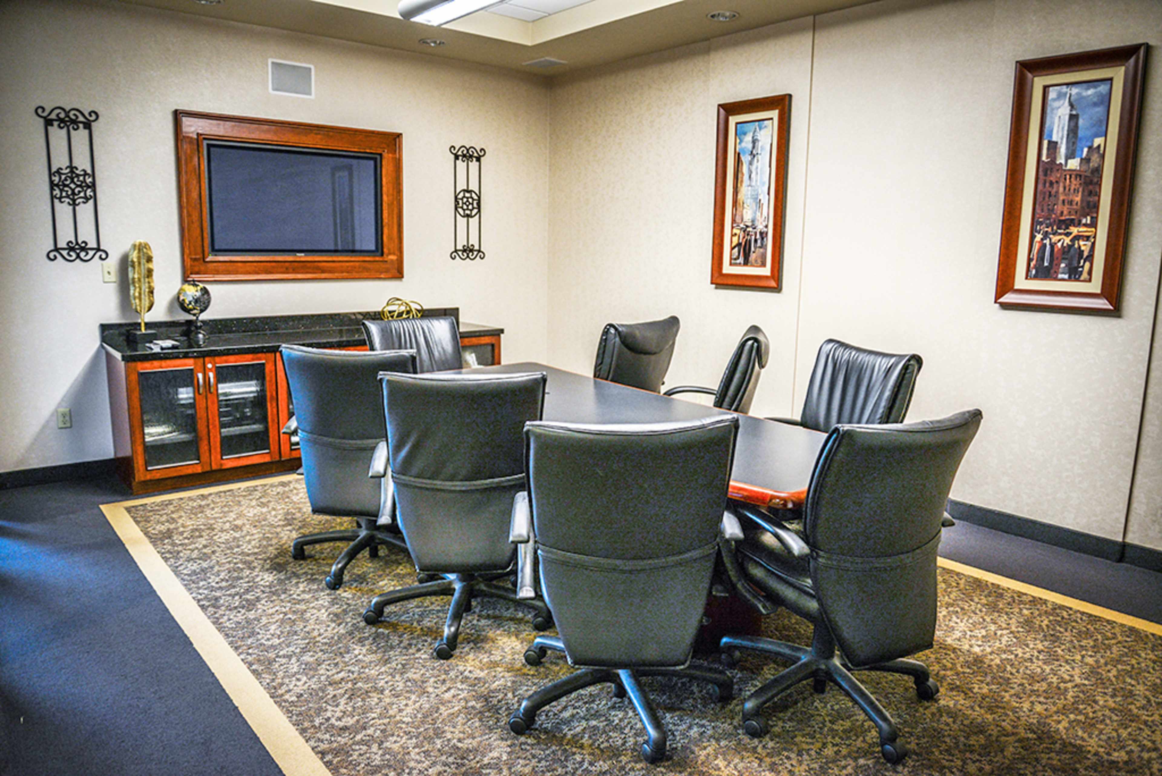 A conference room with a large rectangular table surrounded by black office chairs, two framed pictures on the walls, and a wall-mounted television.