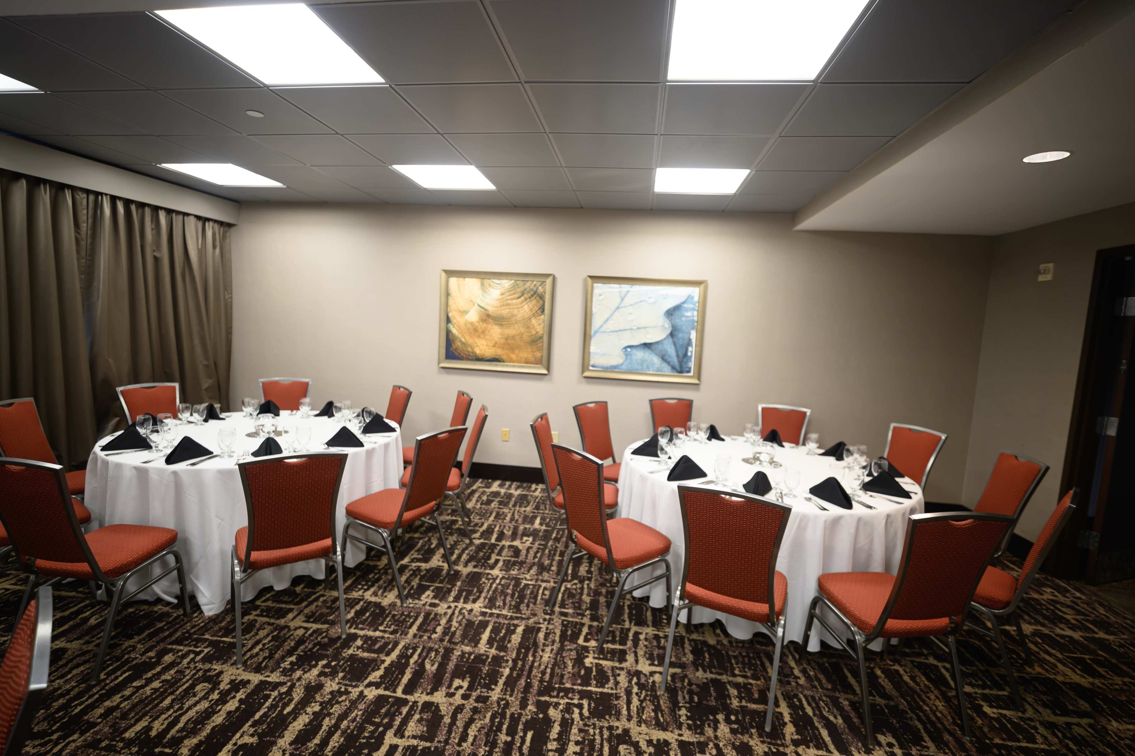 The image shows a meeting room with two round tables set for dinner, each surrounded by red chairs and adorned with white tablecloths and black napkins.