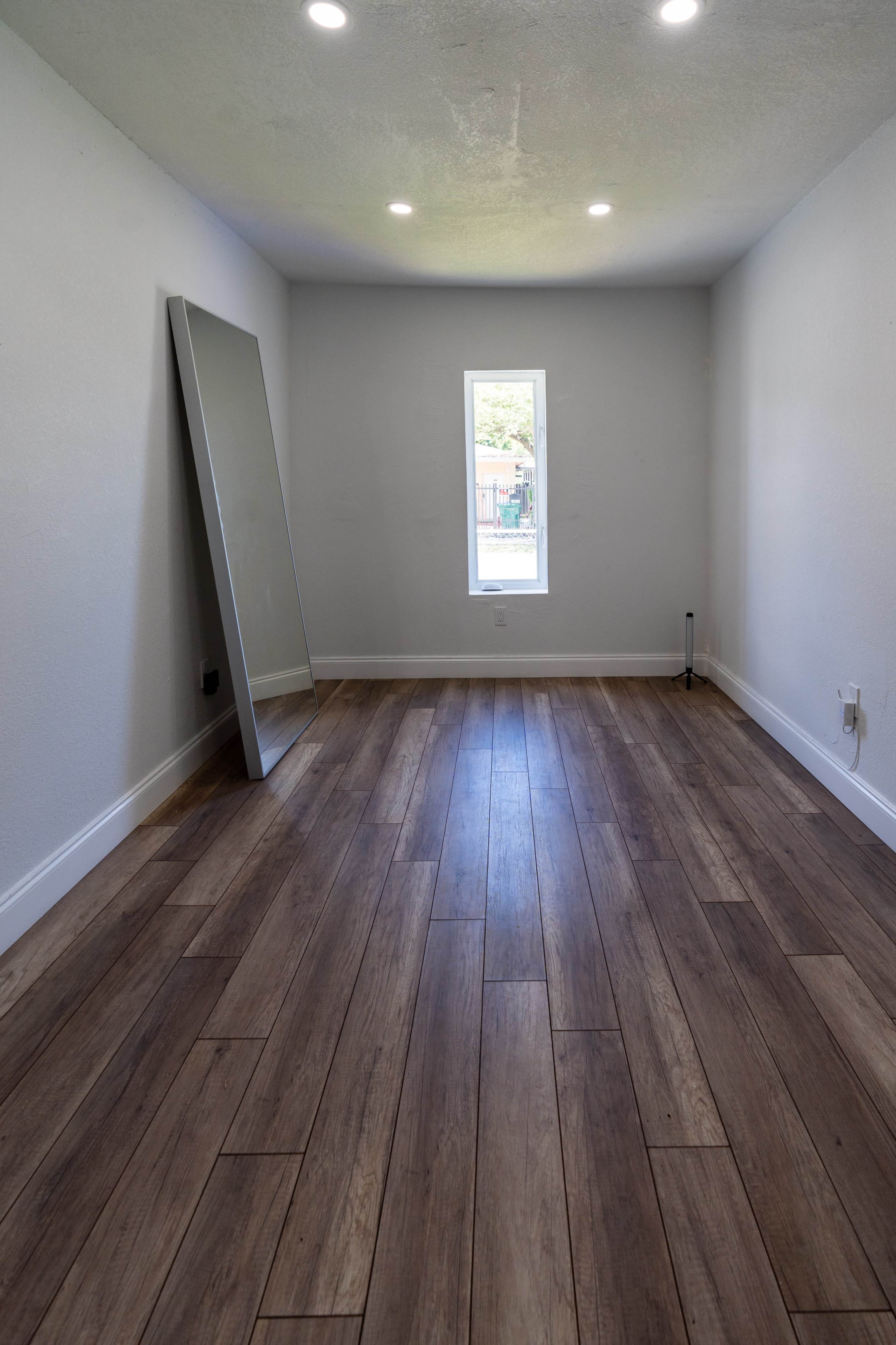 An empty room with wooden flooring, a large mirror leaning against one wall, and a window letting in natural light.
