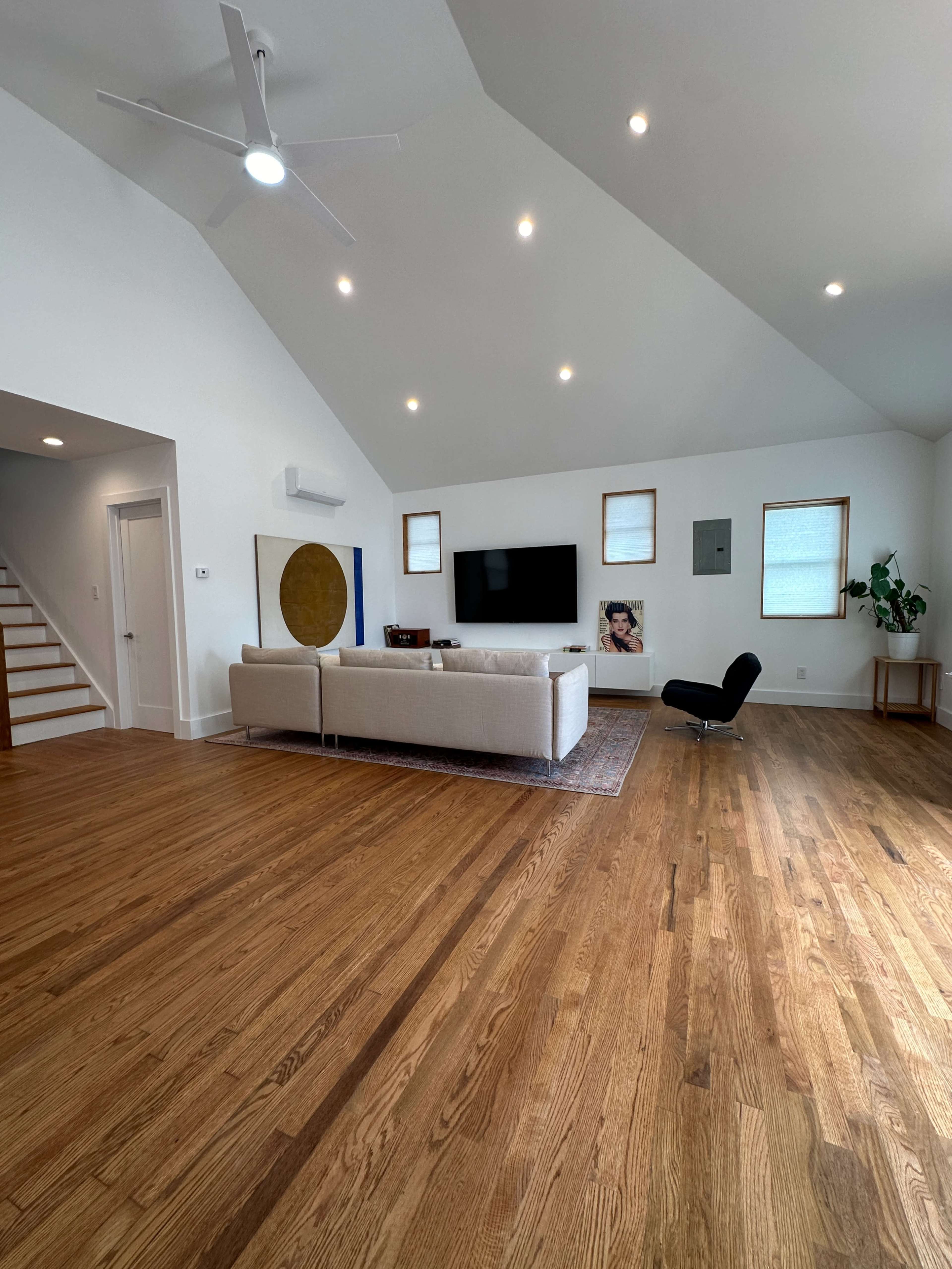 The image shows a modern living room with a light-colored sofa, a television mounted on the wall, and wooden floors, complemented by a staircase and a potted plant.