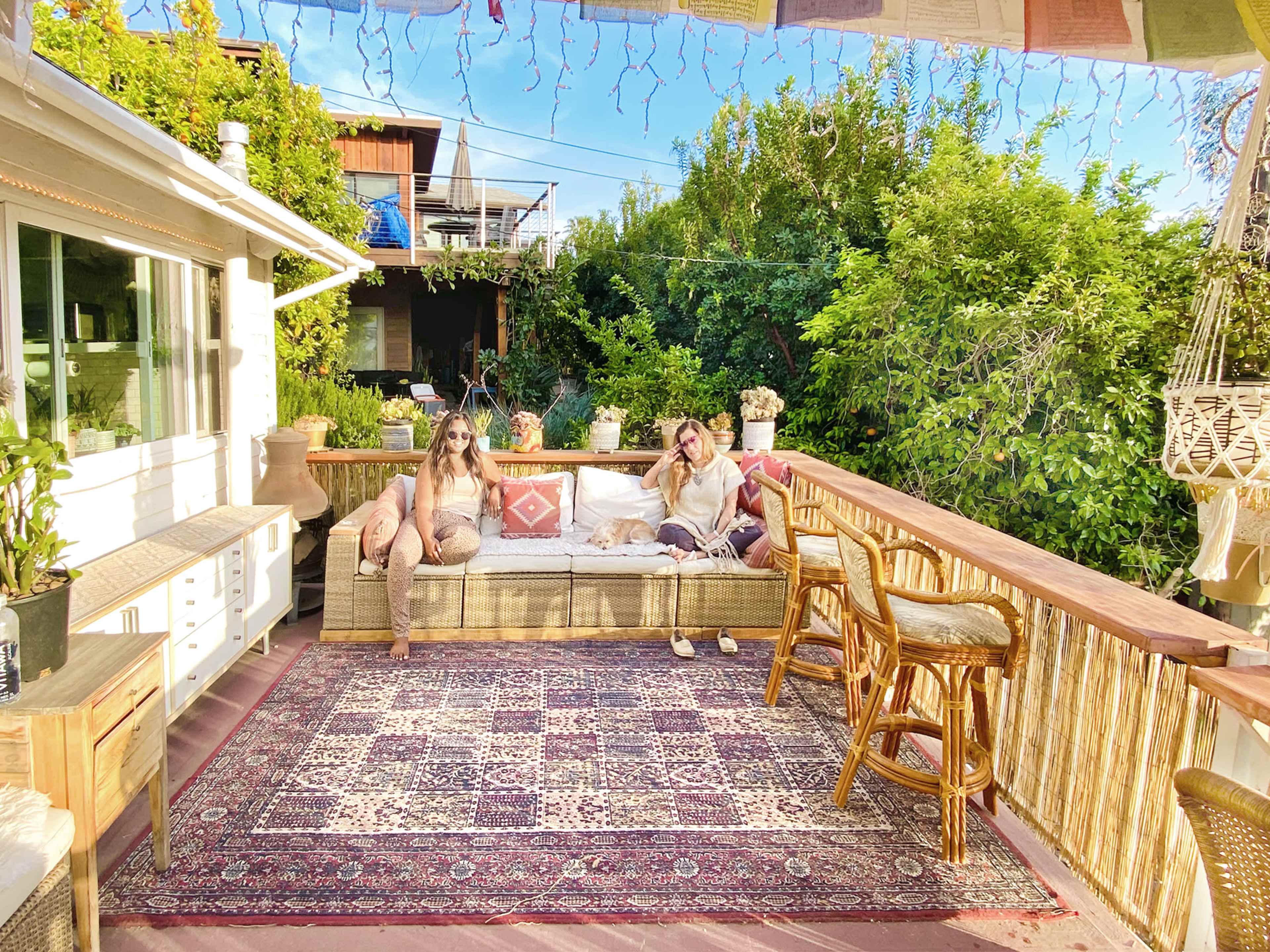 Two women sit on a garden terrace adorned with a woven couch and surrounded by greenery.